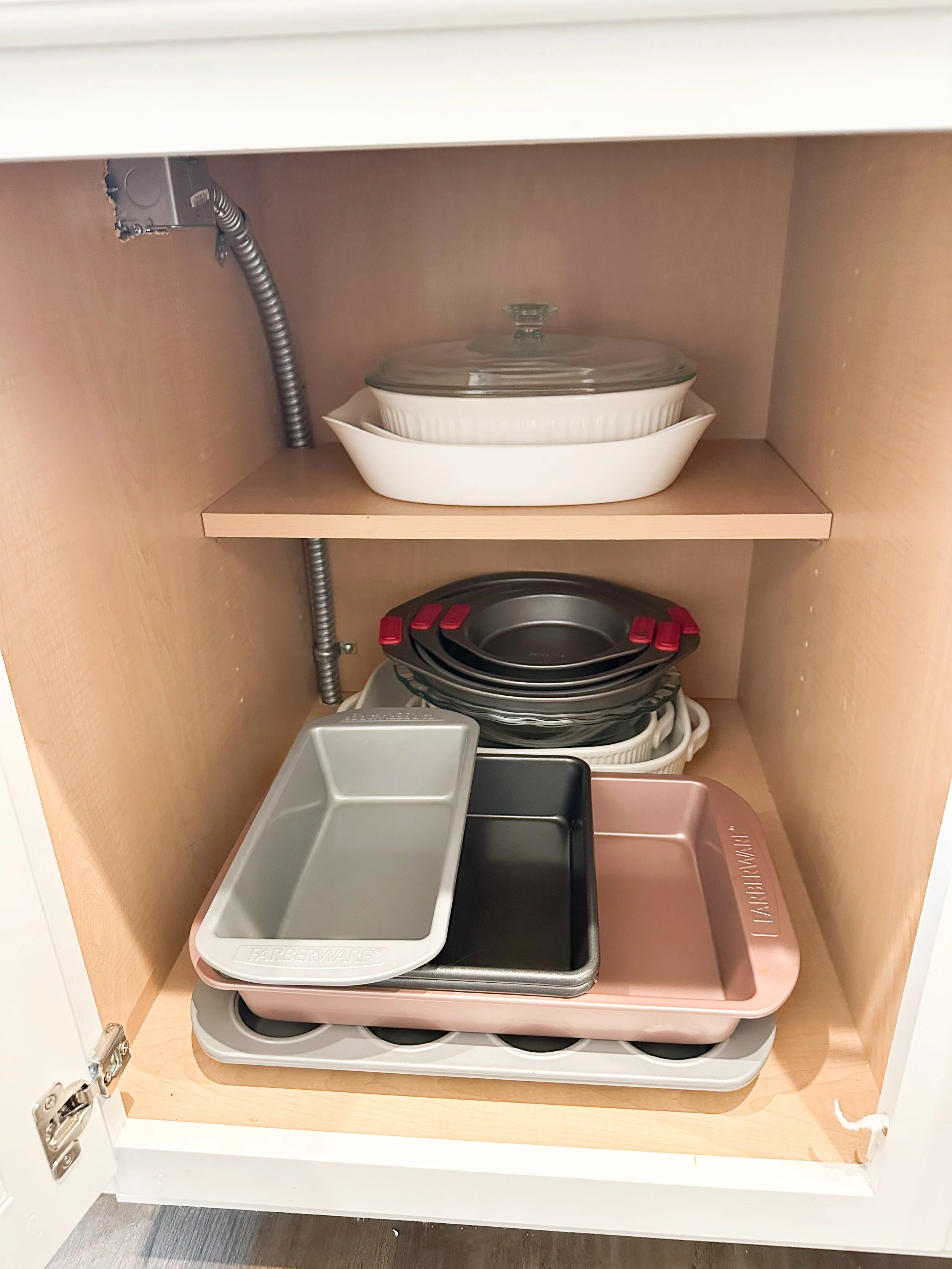 Open kitchen cabinet containing various baking and cooking pans, trays, and dishes stacked inside.