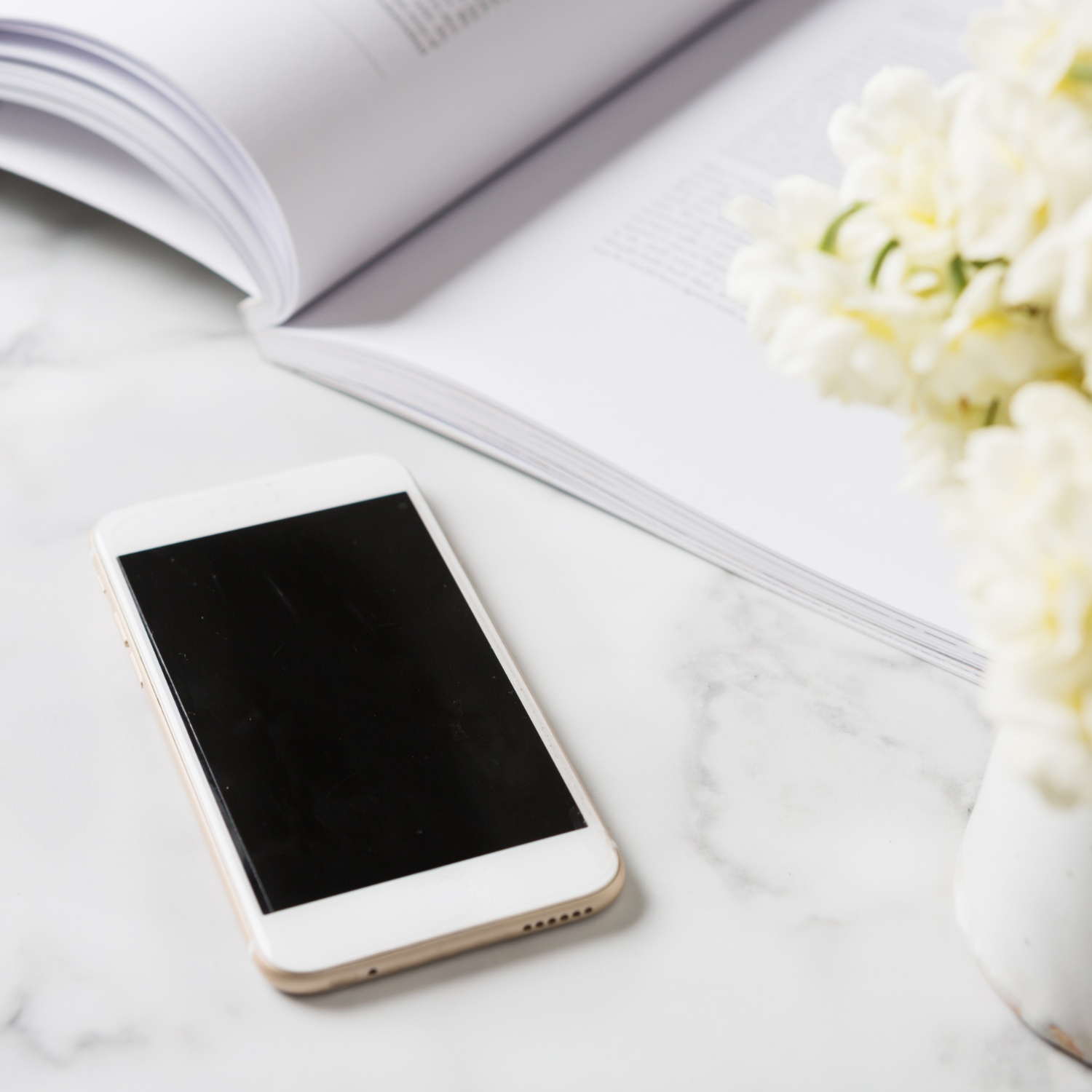 A smartphone with a black screen, an open magazine, and a bouquet of white flowers on a white marble surface.