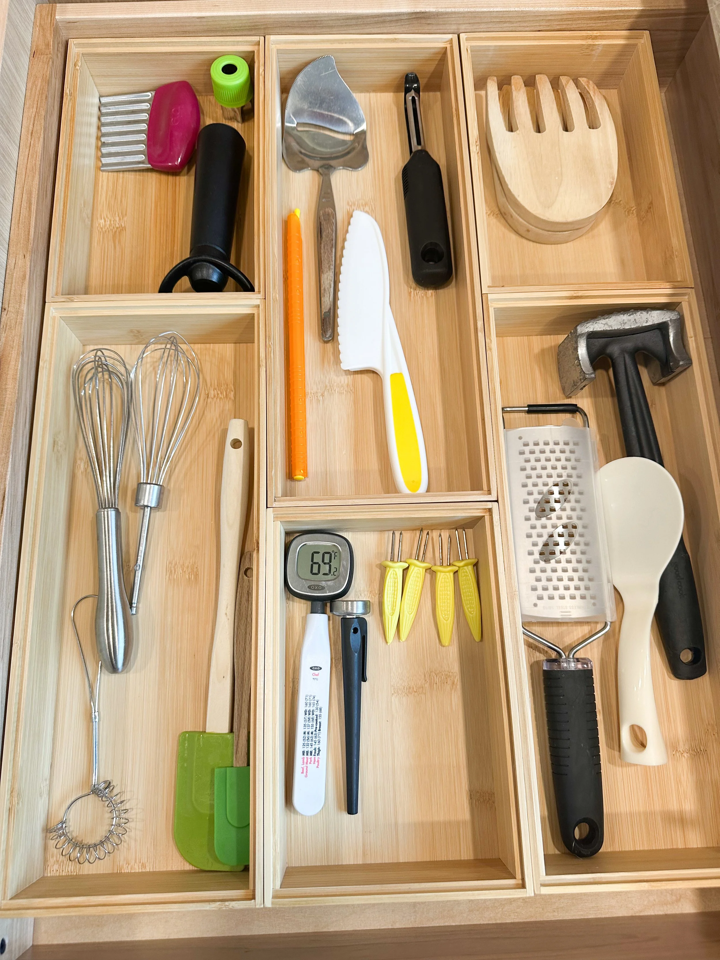 Organization of kitchen tools in wooden compartments including whisks, spatulas, a hammer, small cutters, a scraper, a grater, a thermometer, and small kitchen gadgets.