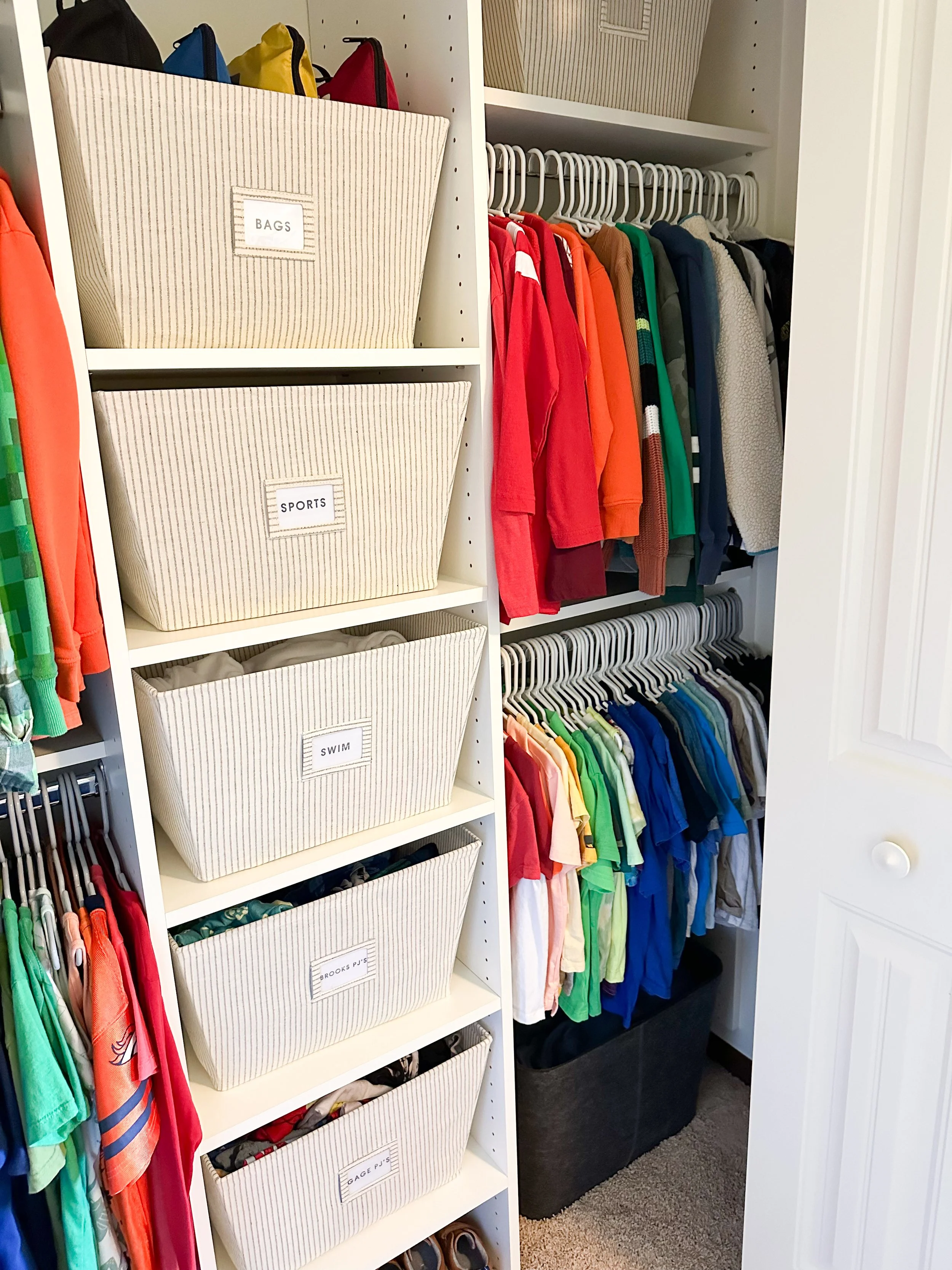 Closet with hanging colorful children's clothes and labeled storage bins on a white shelf for bags, sports gear, swimwear, pajamas, and other clothing.