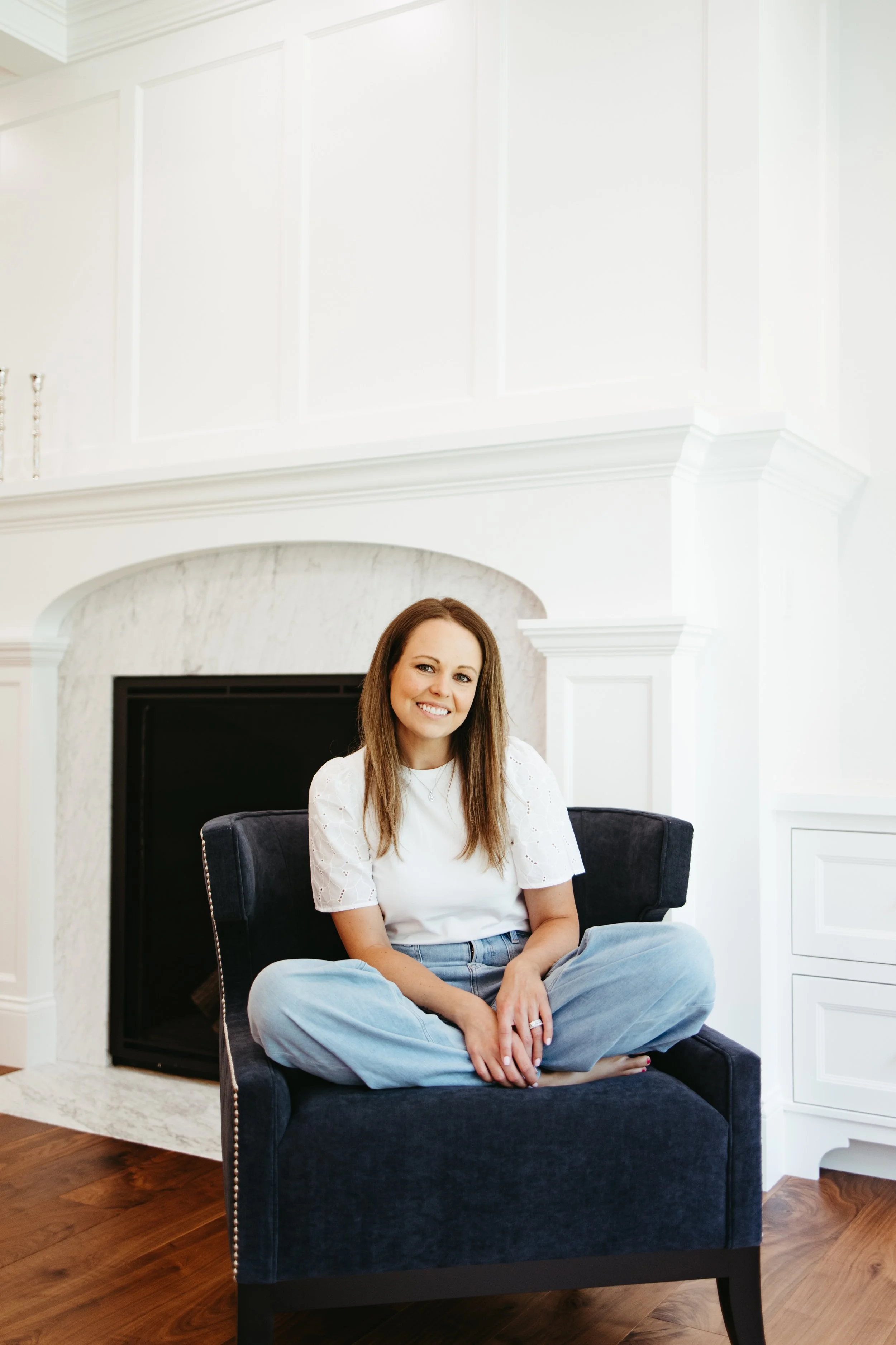 A woman with long brown hair, wearing a white shirt and light blue jeans, sitting cross-legged on a dark blue armchair in a bright, modern living room with white walls and wooden flooring, smiling at the camera.