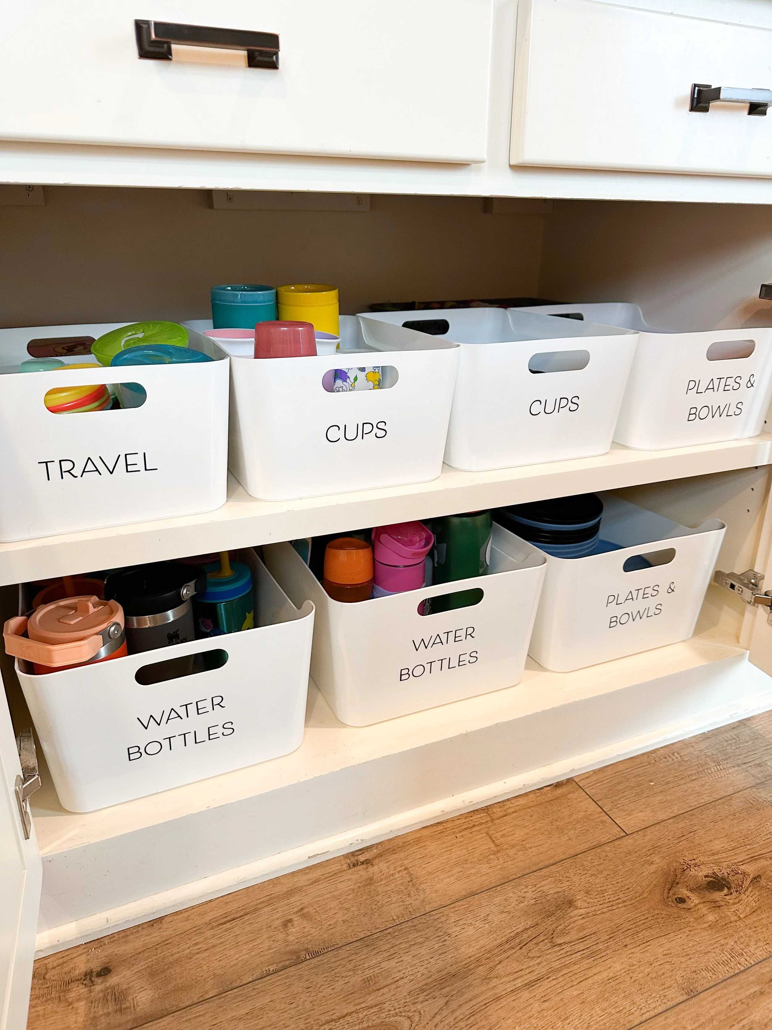 Open kitchen cabinet with labeled white baskets containing cups, plates, bowls, and water bottles.