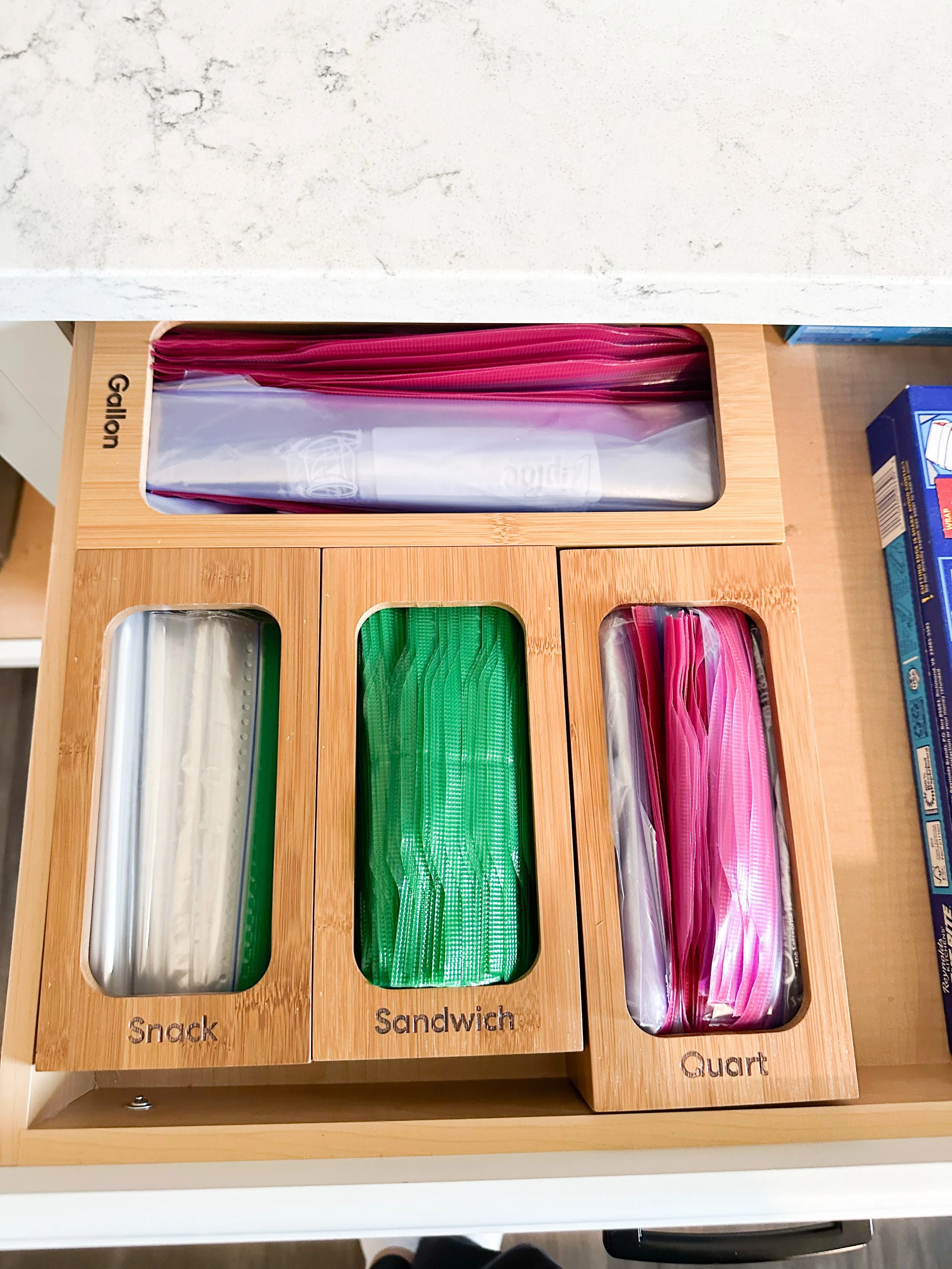 A wooden organizer with labeled compartments containing plastic sandwich, snack, and quart-sized zipper bags.