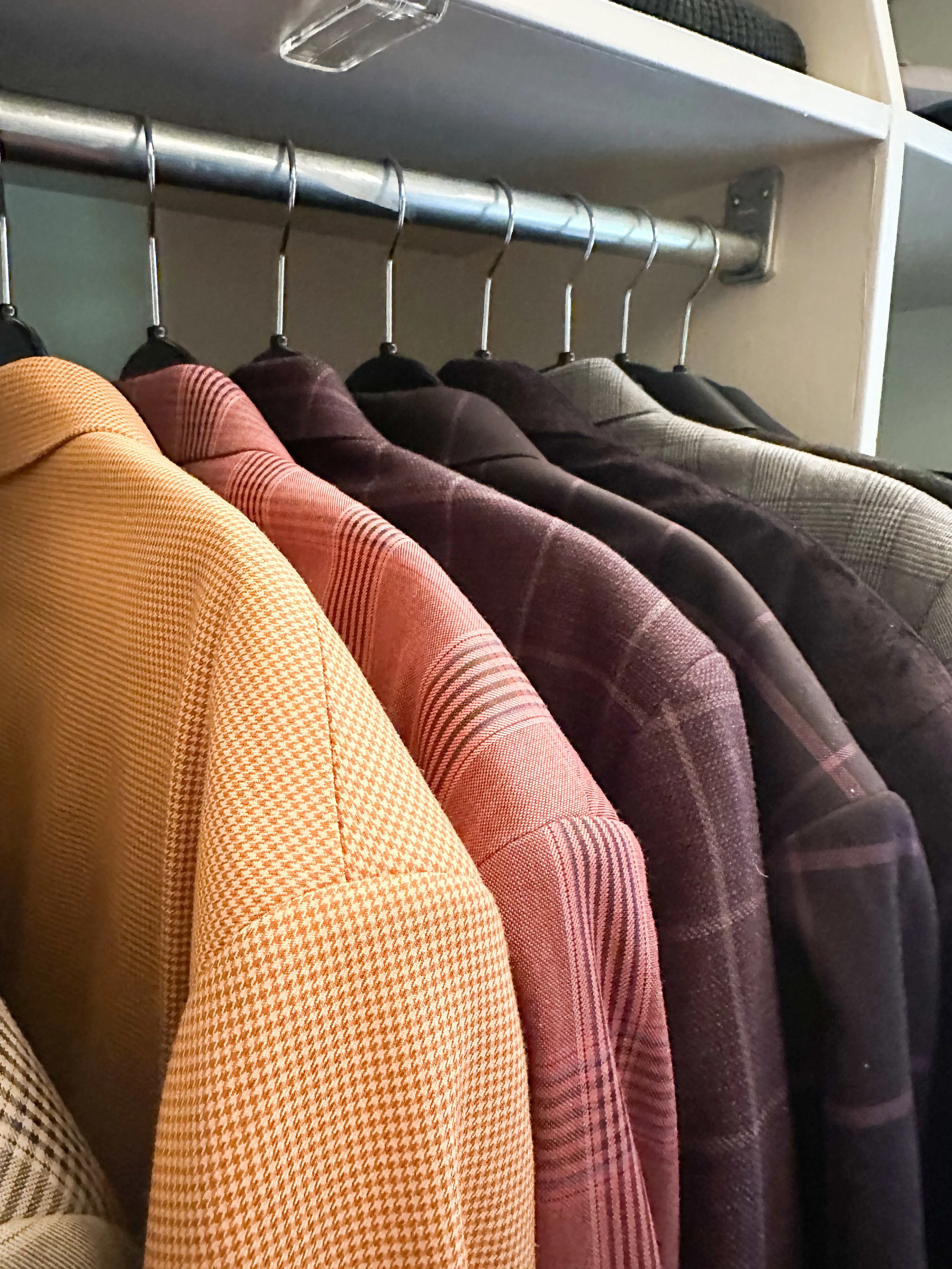 A row of men's blazers hanging on a closet rod, featuring various plaid and checkered patterns in shades of yellow, pink, red, brown, and gray.