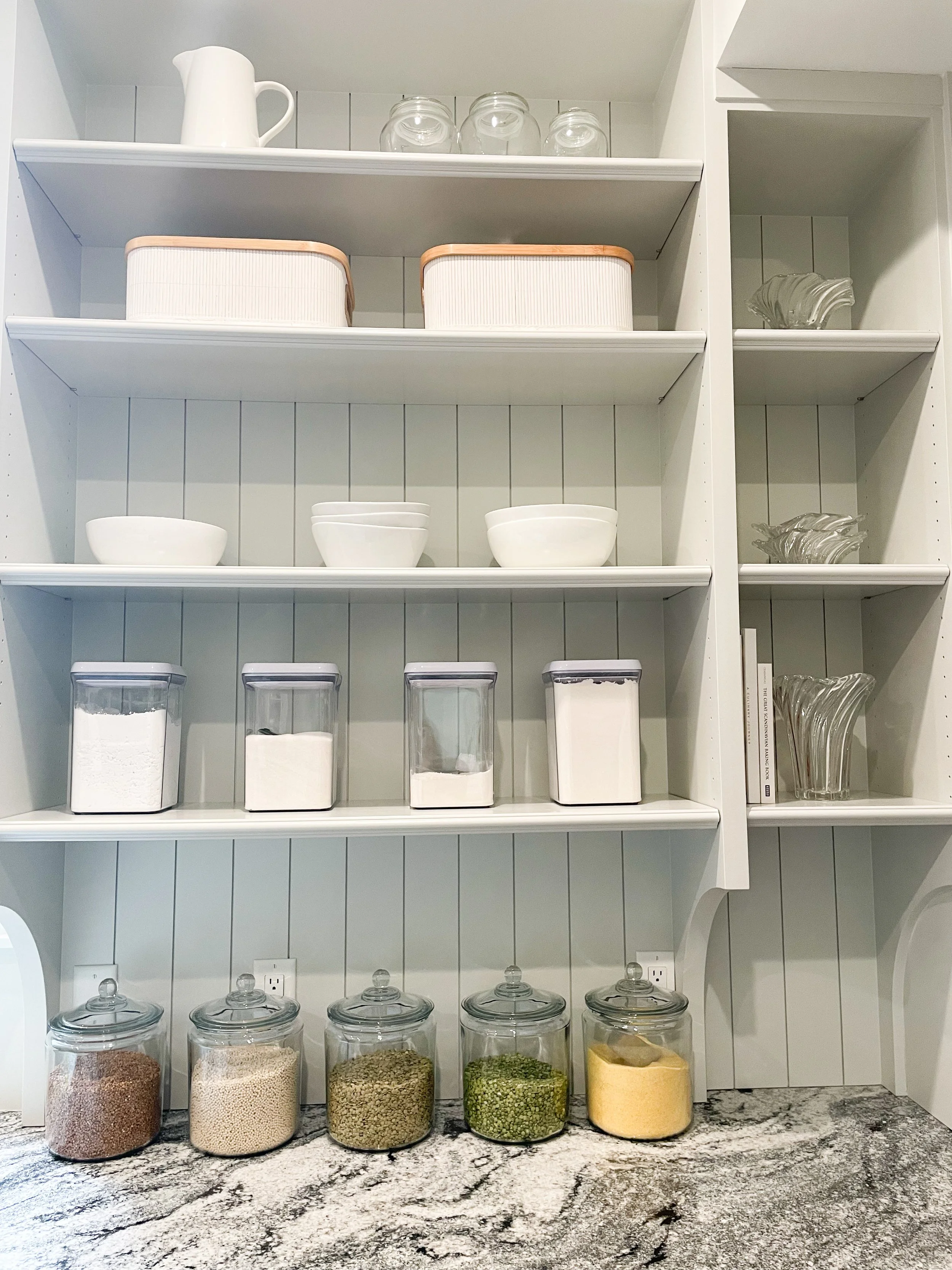 Open white kitchen shelves with jars of spices, bowls, and glassware, and a granite countertop.