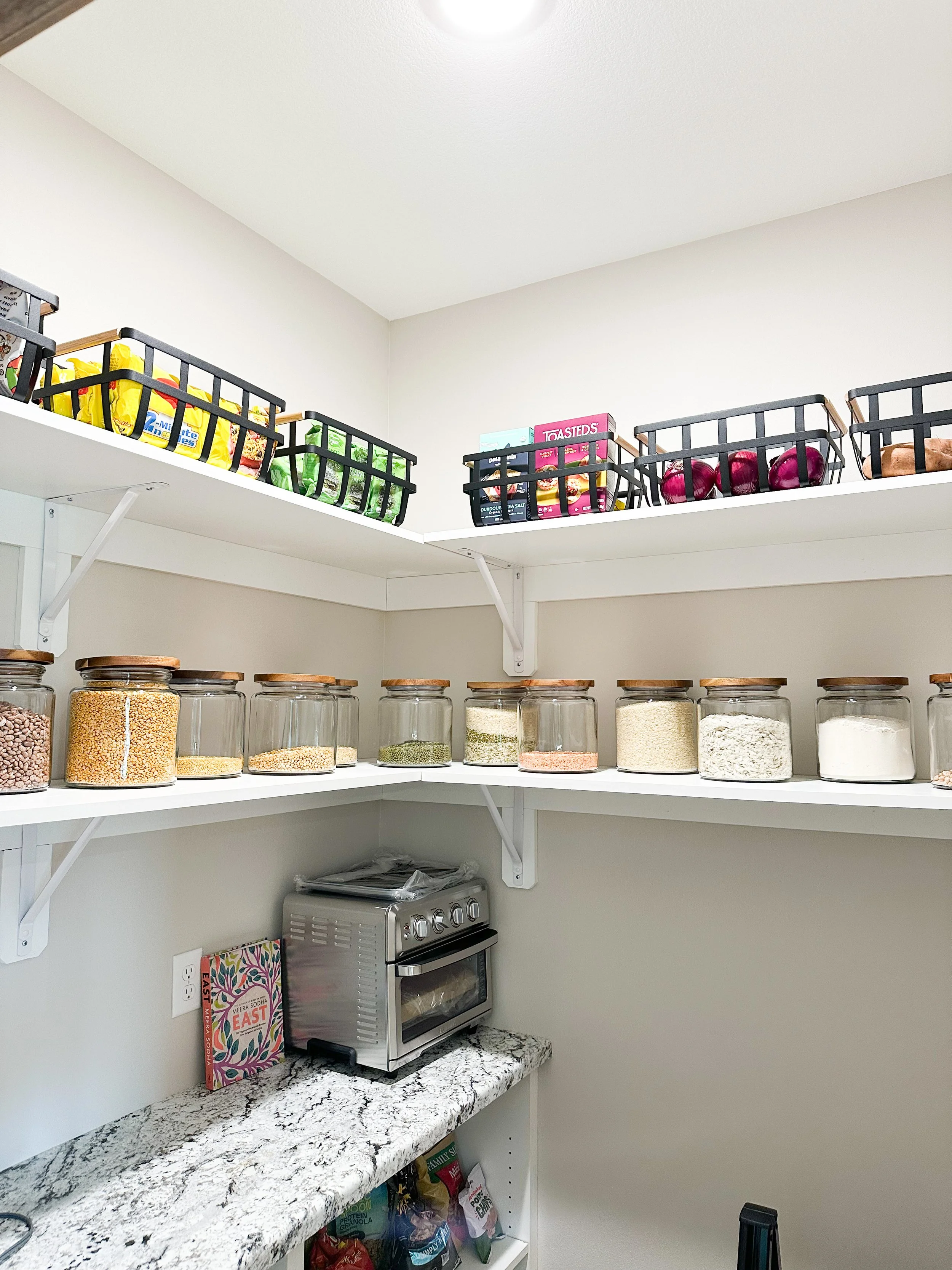 Pantry with glass jars of grains and spices on white shelves, black baskets with food items on top, small oven on granite countertop, and a cookbook beside it.