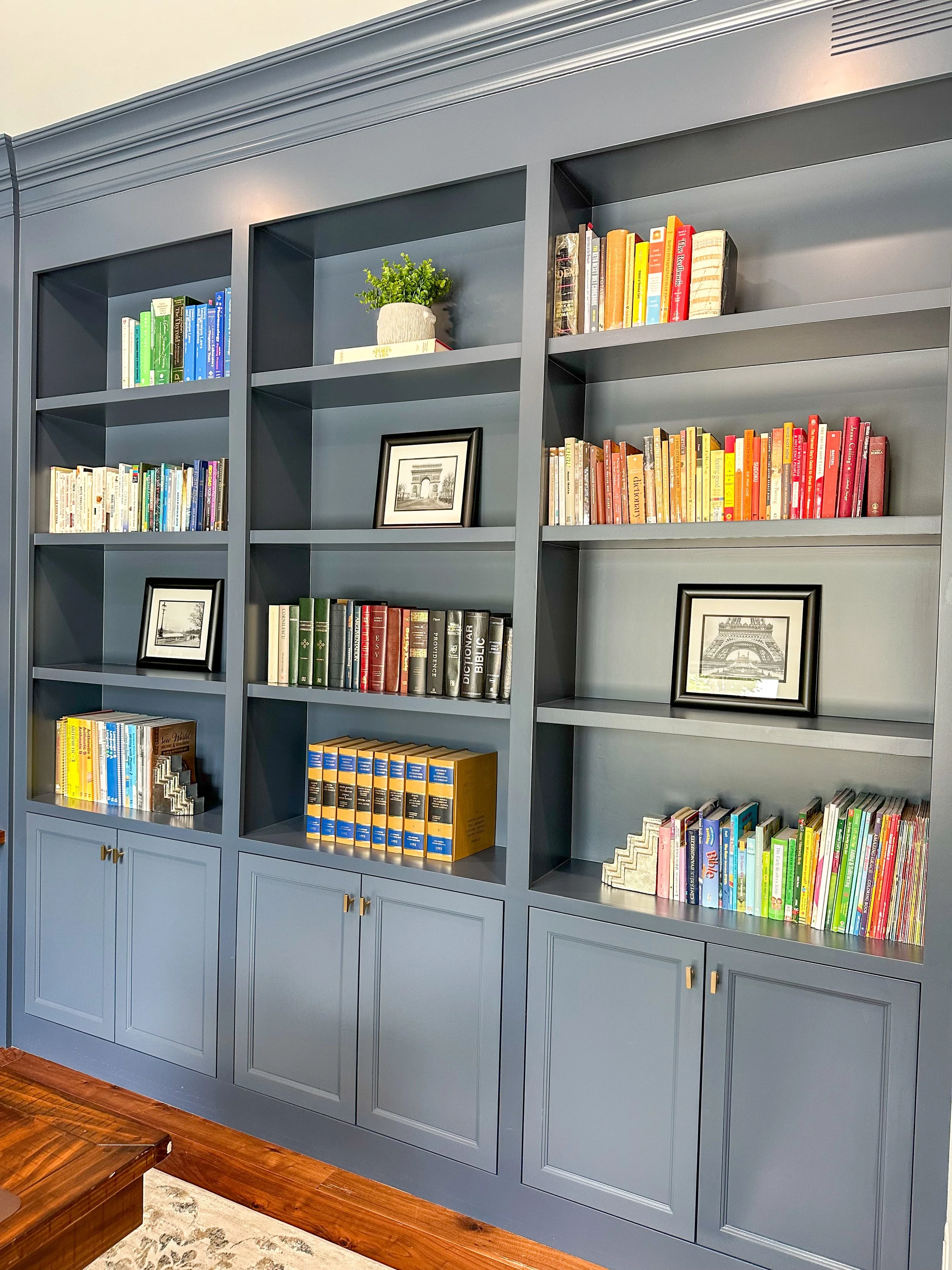 A blue bookshelf with multiple books, framed pictures, a potted plant, and decorative items in a living room.