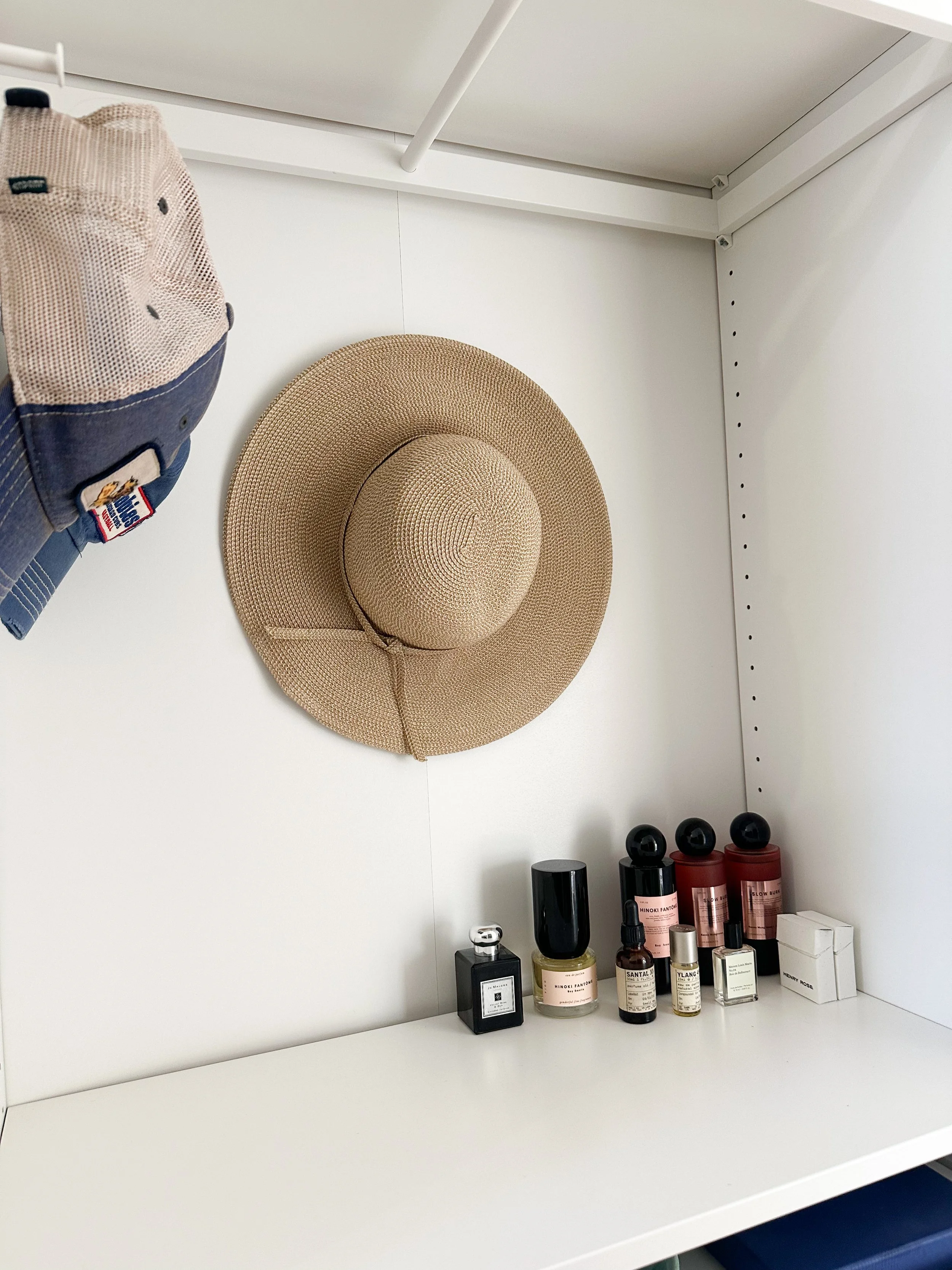A beige straw wide-brimmed hat hanging on a white wall, beside various perfume bottles and a small white box on a white shelf.