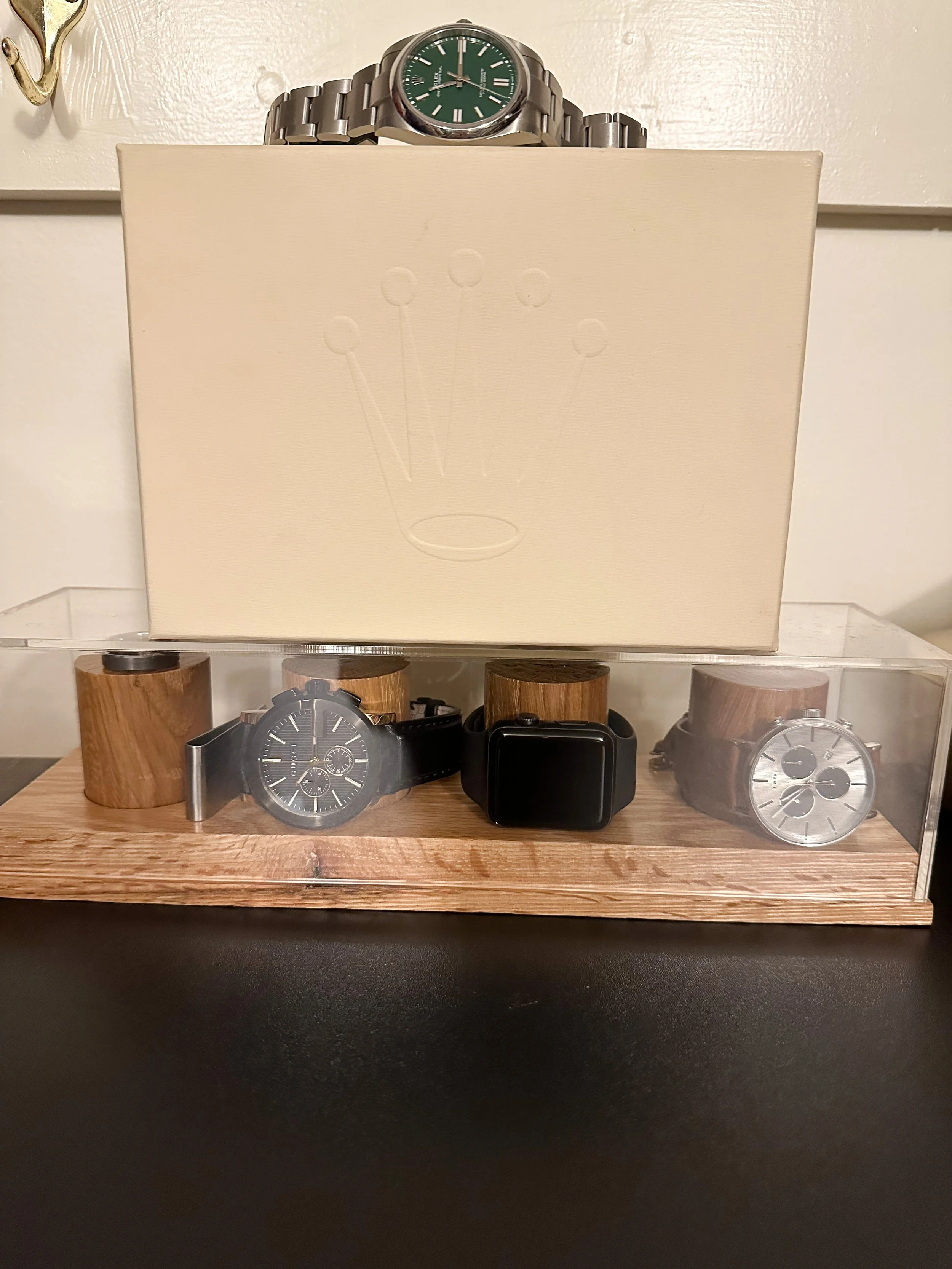 Three wristwatches on wooden display stands inside a clear plastic case, with a boxed watch on top of the display. The watches include a black chronograph, a black smartwatch, and a silver chronograph. The box features a subtly embossed crown logo.