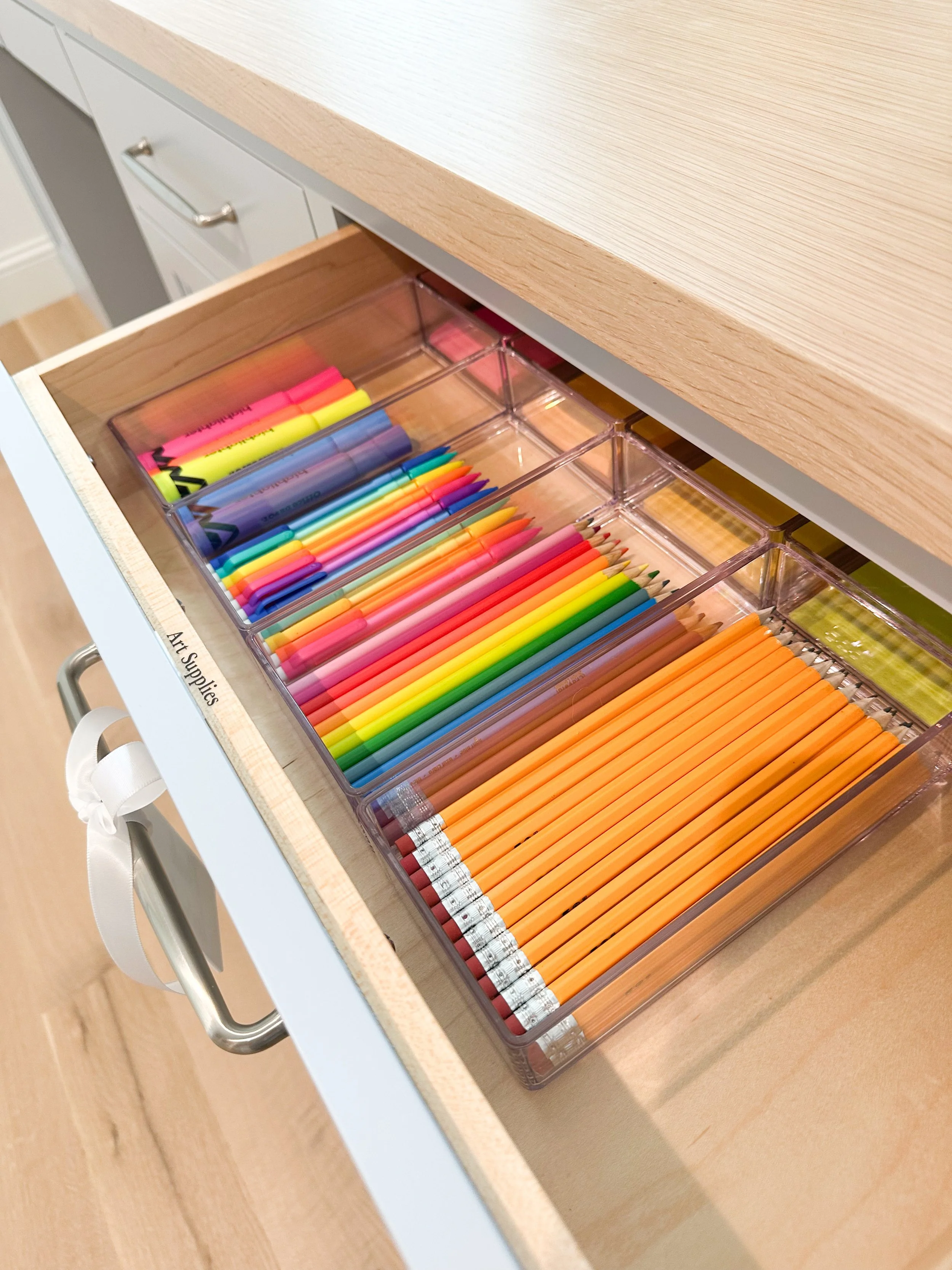 Open drawer with labeled compartments holding colorful highlighters, markers, and pencils inside a wooden cabinet.