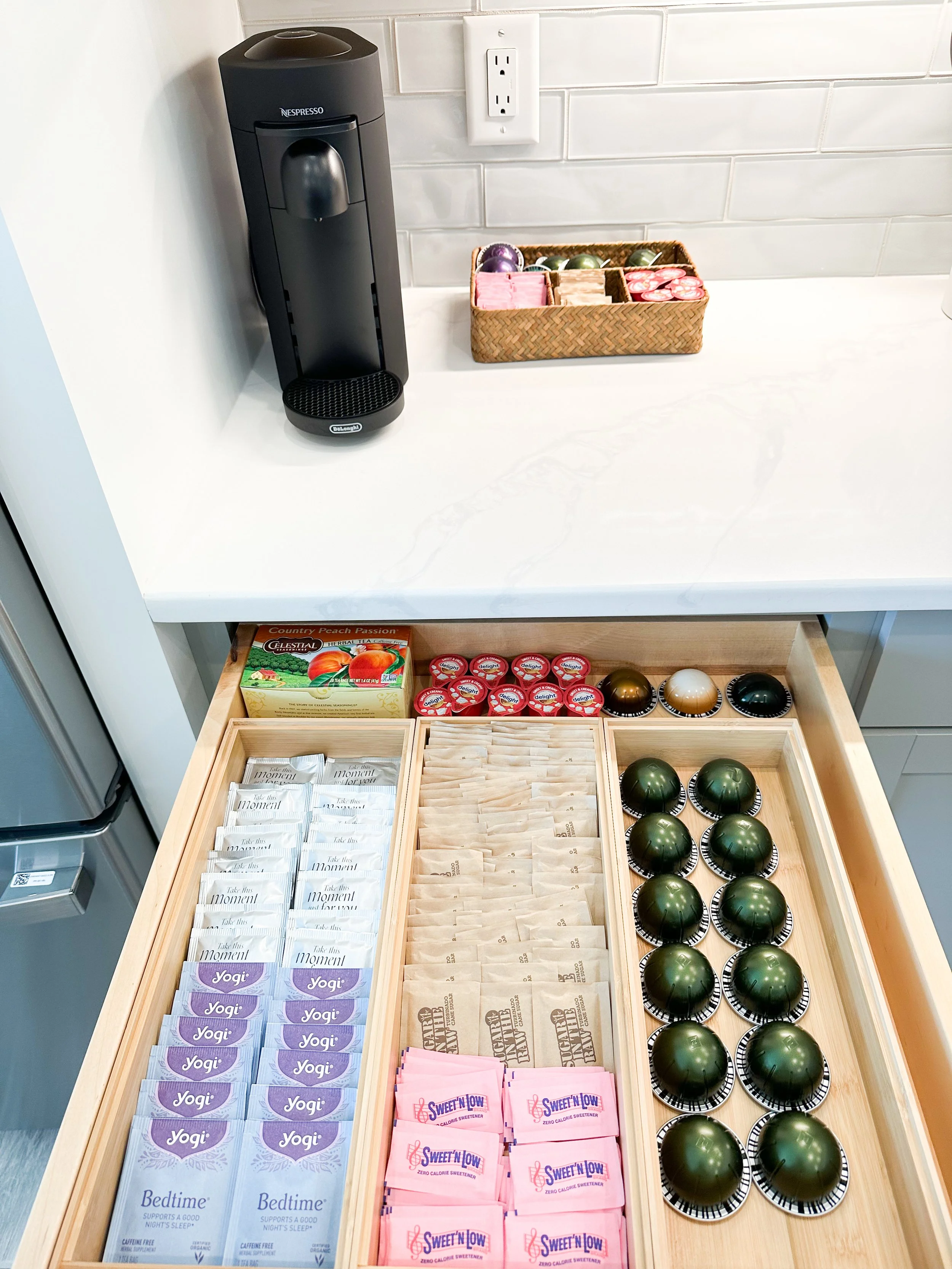 A countertop with a Nespresso coffee machine, a woven basket with tea and coffee pods, and an open drawer filled with various coffee and tea packet options, including caffeine-free, yogi, sweet'n low, and different colored coffee capsules.