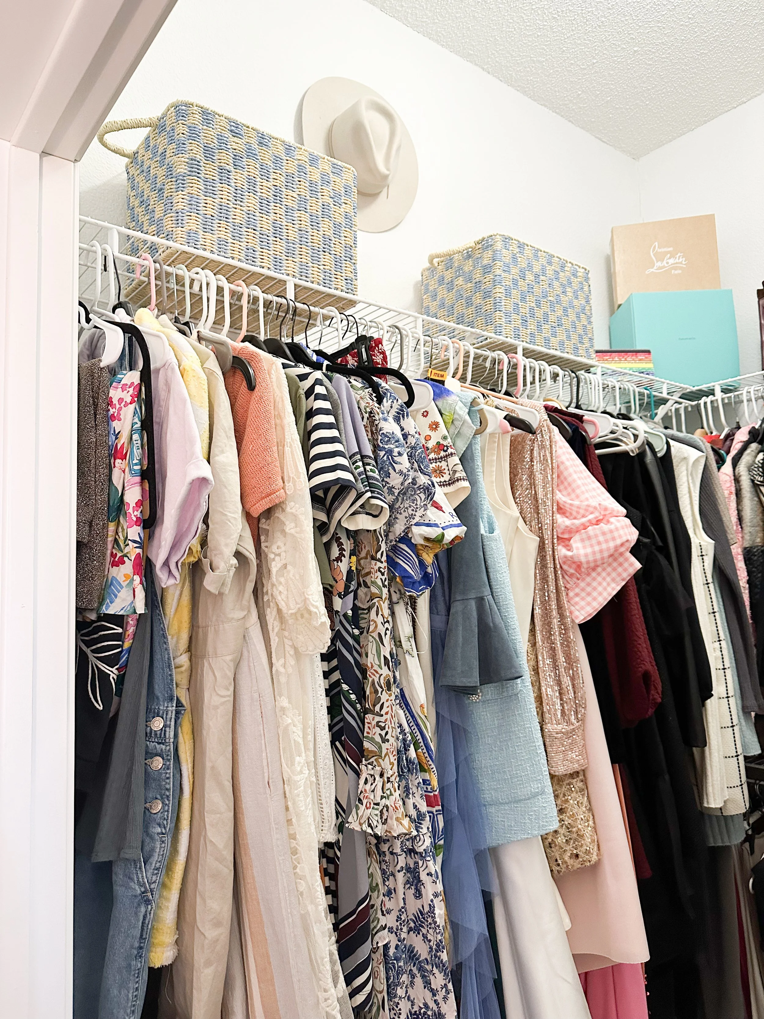 A closet with a variety of hanging clothes including dresses, shirts, and jackets. Top shelf with woven baskets and a box, wall mounted hat, and a few shoe boxes.