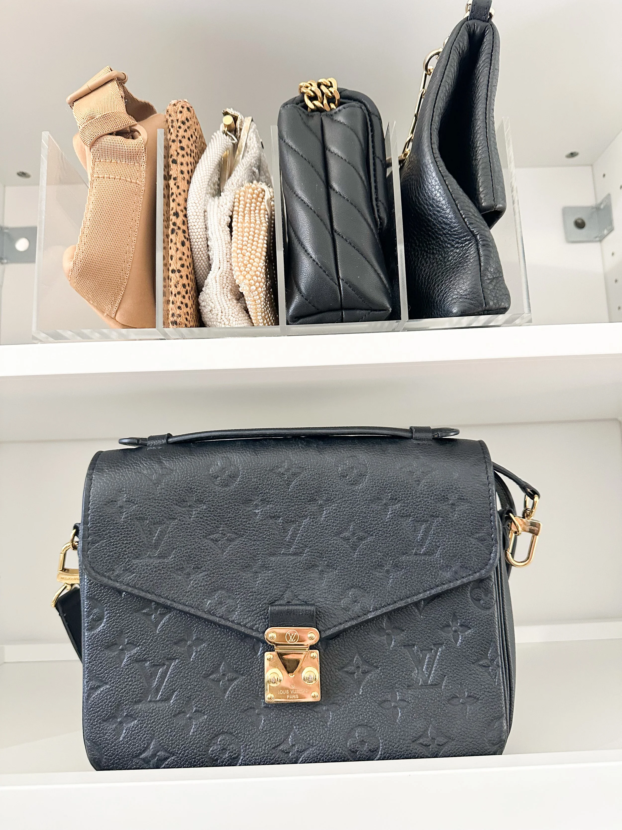 A black Louis Vuitton handbag with a gold clasp on a white shelf, with a clear plastic container holding several other handbags and pouches above it.