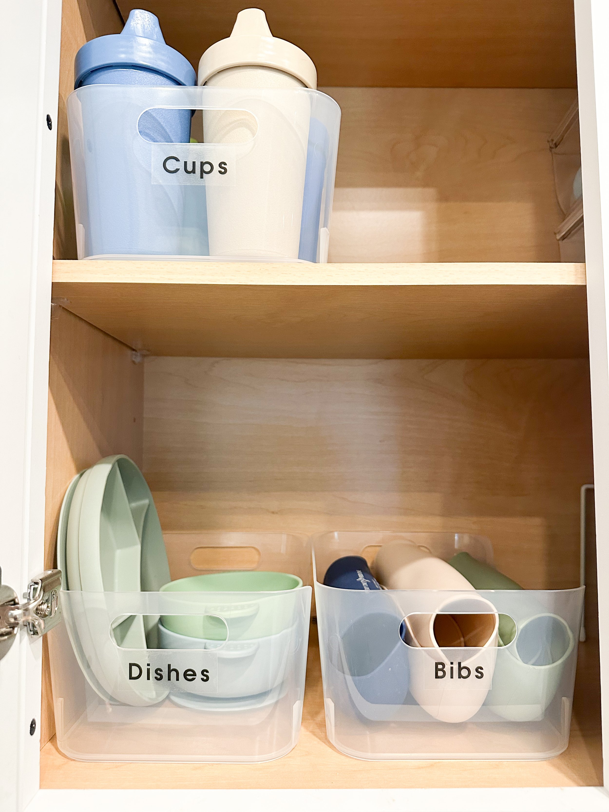 Kitchen cabinet with plastic storage bins labeled 'Cups', 'Dishes', and 'Bibs'. The 'Cups' bin contains two sippy cups, and the 'Dishes' bin holds bowls and plates, while the 'Bibs' bin contains bibs and cups.