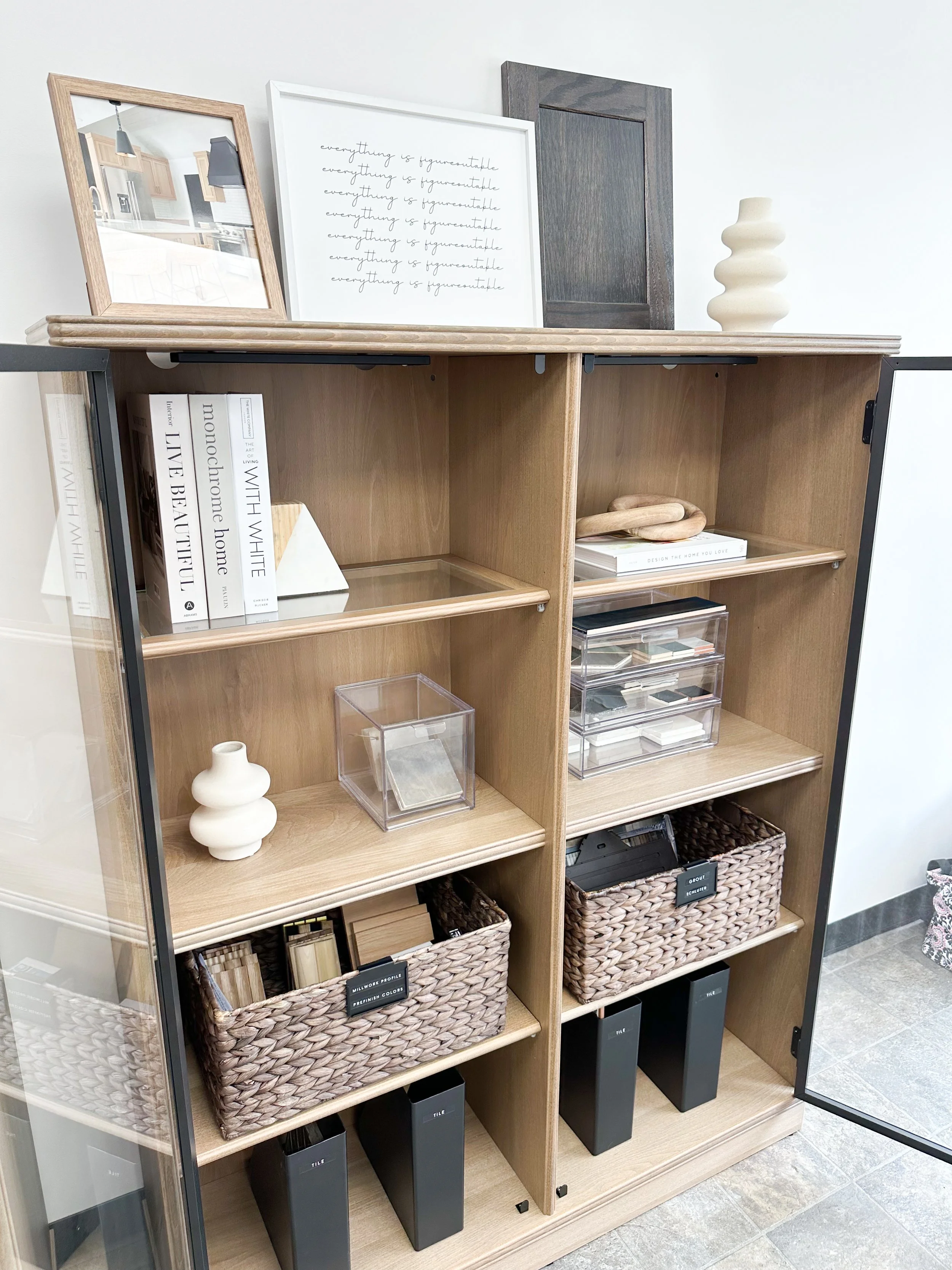 A wooden cabinet with glass doors and multiple shelves, holding decorative objects, books, storage boxes, and black containers, with framed artwork and a vase on top.