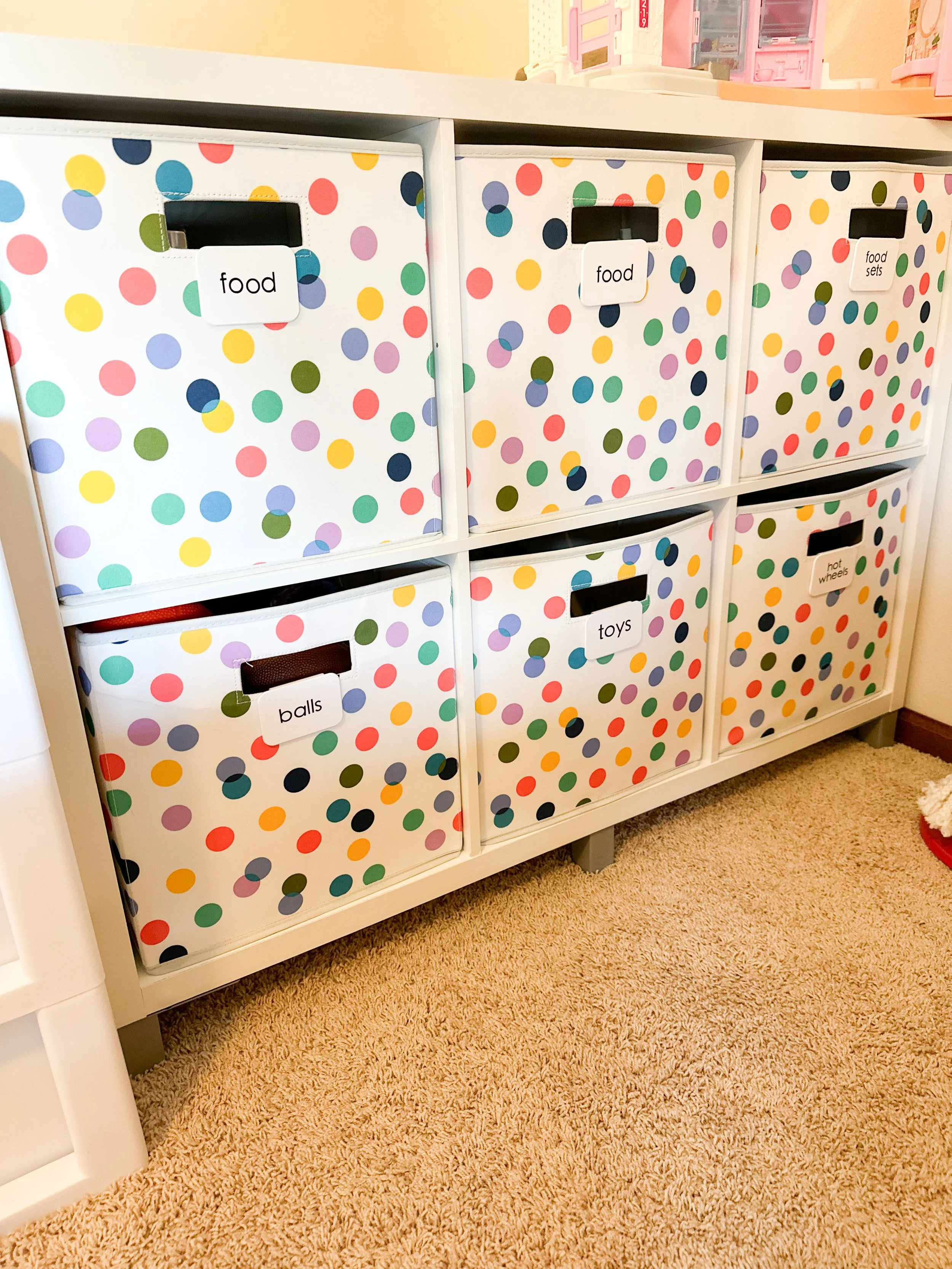 Colorful polka dot storage bins labeled with various household categories like food, toys, balls, hot wheels, and food sets, arranged in a white shelving unit on a carpeted floor.