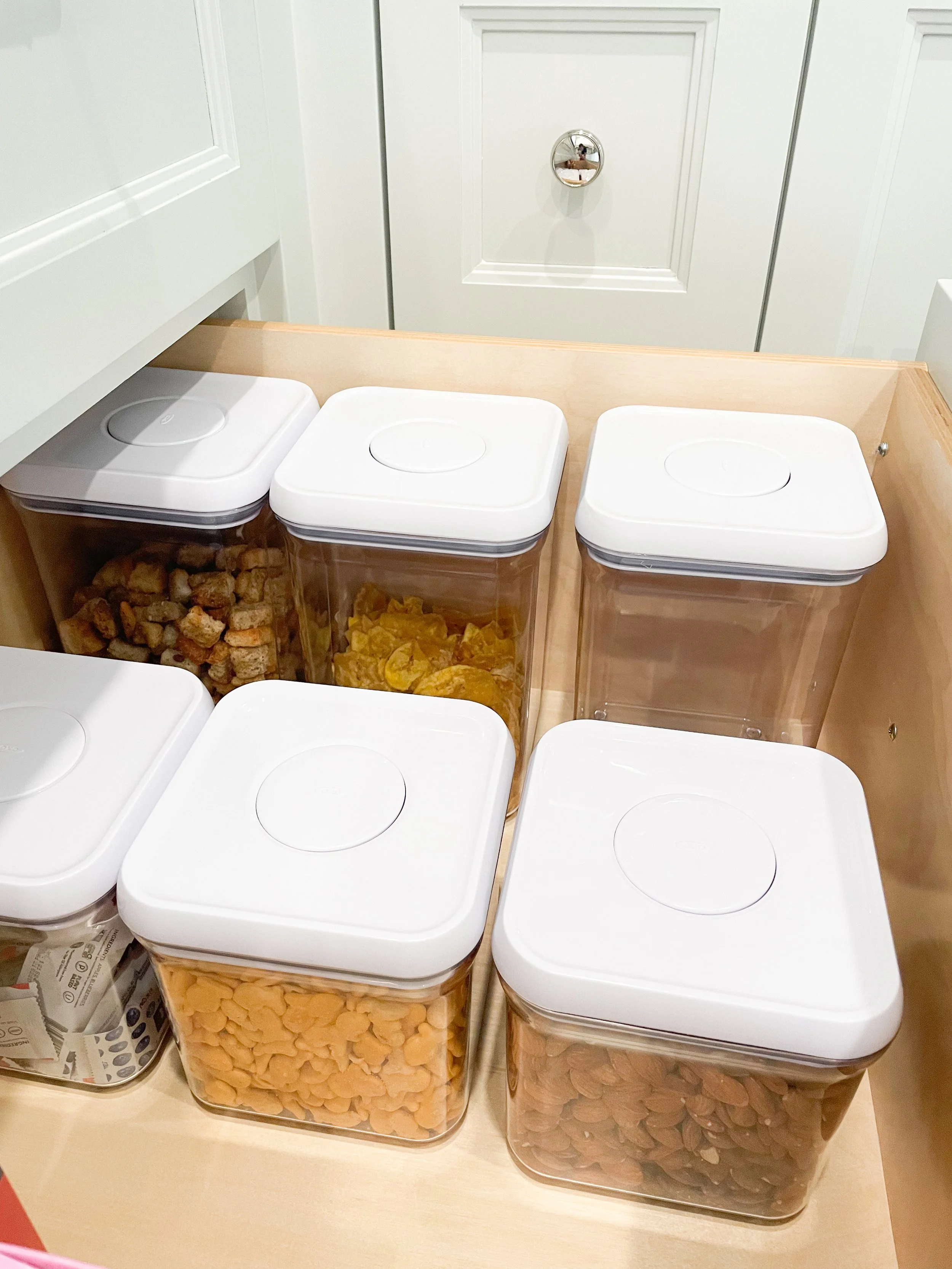 Six transparent storage bins with white lids, containing snacks such as candy, chips, and nuts, arranged on a light wood surface in a kitchen or pantry.