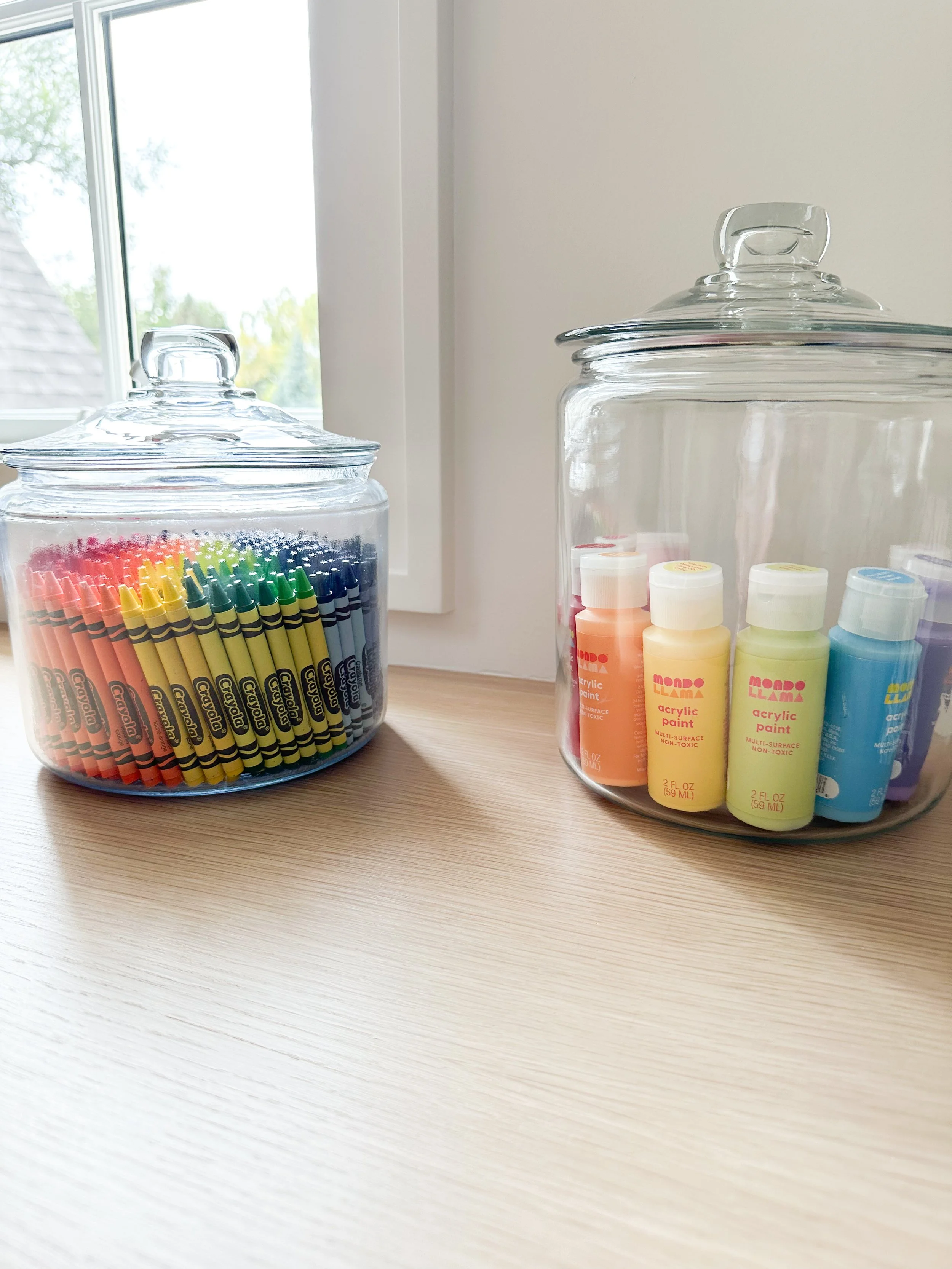 Two glass jars with colorful markers and paint bottles on a wooden surface by a window.
