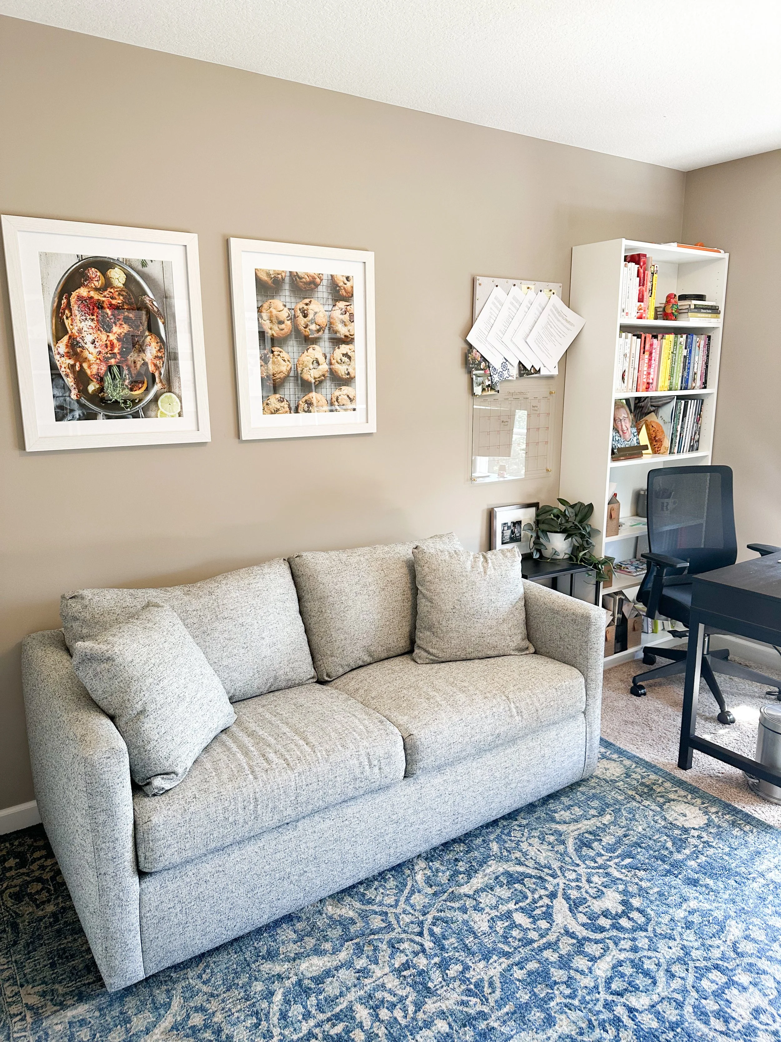 Living room with a beige sofa, navy blue area rug, white bookshelf filled with books and decor, black desk with an office chair, framed food photos on the wall, small side table with a plant, and papers pinned to a wall organizer.