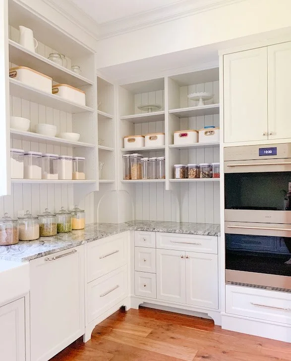A white kitchen with open shelving, glass jars filled with grains and spices, white bowls, and storage containers, granite countertops, and built-in oven and microwave.