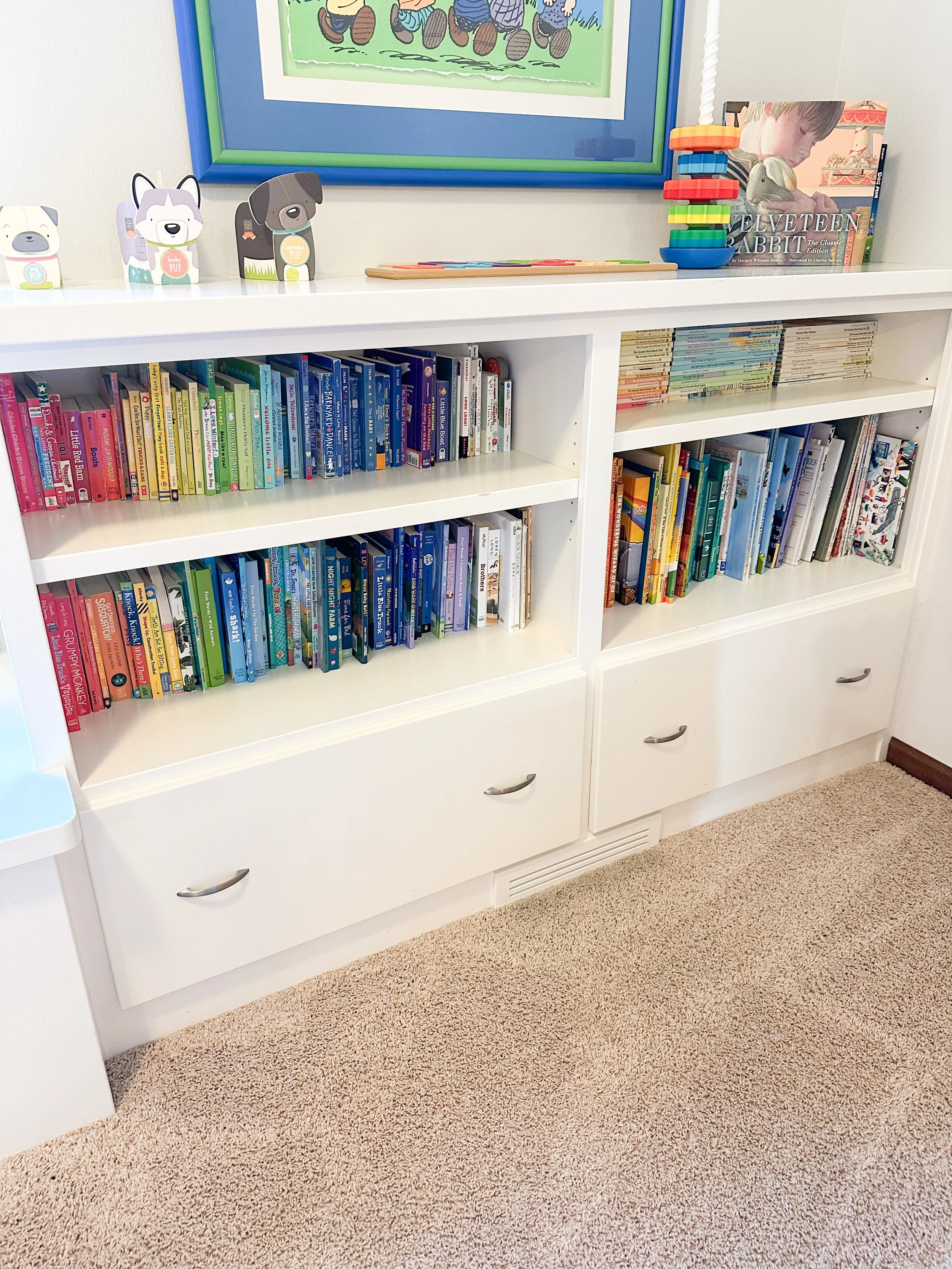 White bookshelf with three shelves filled with children's books arranged by color, with three drawers at the bottom, a pile of colorful stacking rings, and children’s book illustrations on the wall above.