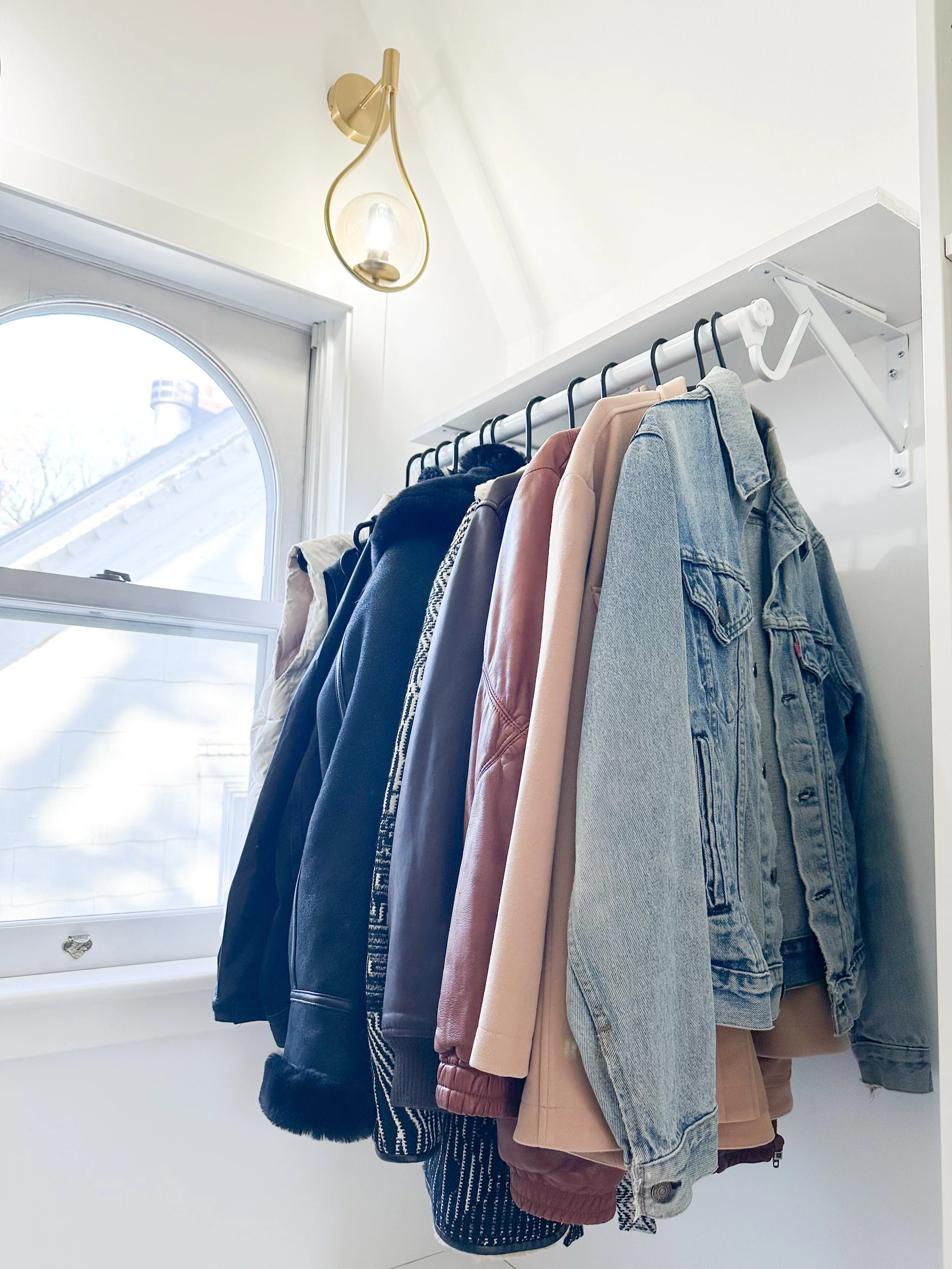 Clothes hanging on a white wall shelf above a window, including jackets, coats, and shirts in various colors and materials.