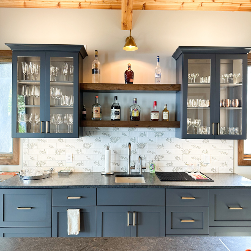 Modern kitchen with blue cabinets, glass-front upper cabinets, and a wooden shelf with liquor bottles, a stainless steel sink, and a stove.