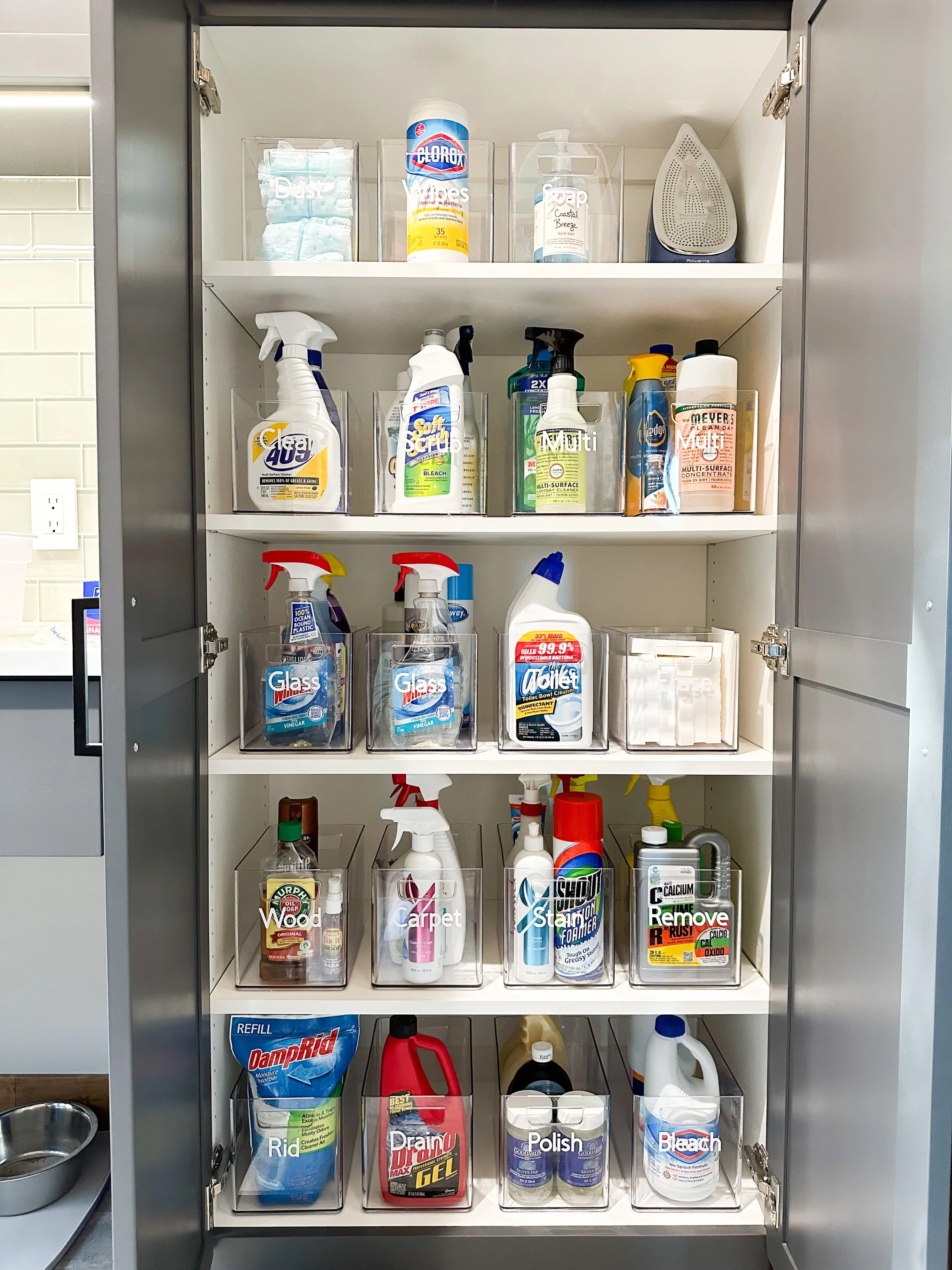 A cabinet filled with various cleaning supplies, including sprays, sprays bottles, bleach, and other cleaning products organized in clear containers.