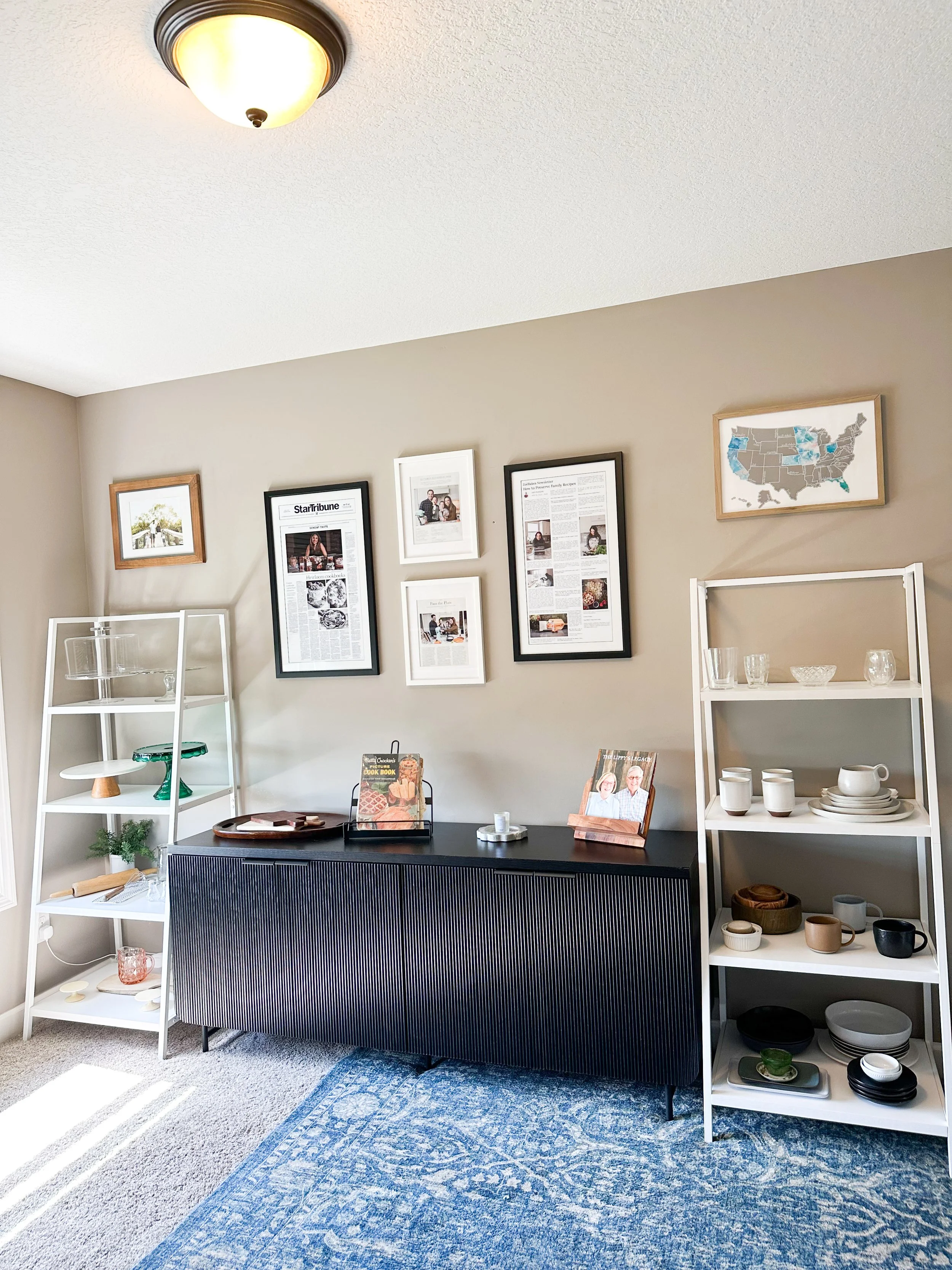 Living room with beige walls, a black sideboard, white shelves with dishes and glassware, framed pictures and a map on the wall, and a blue patterned rug.