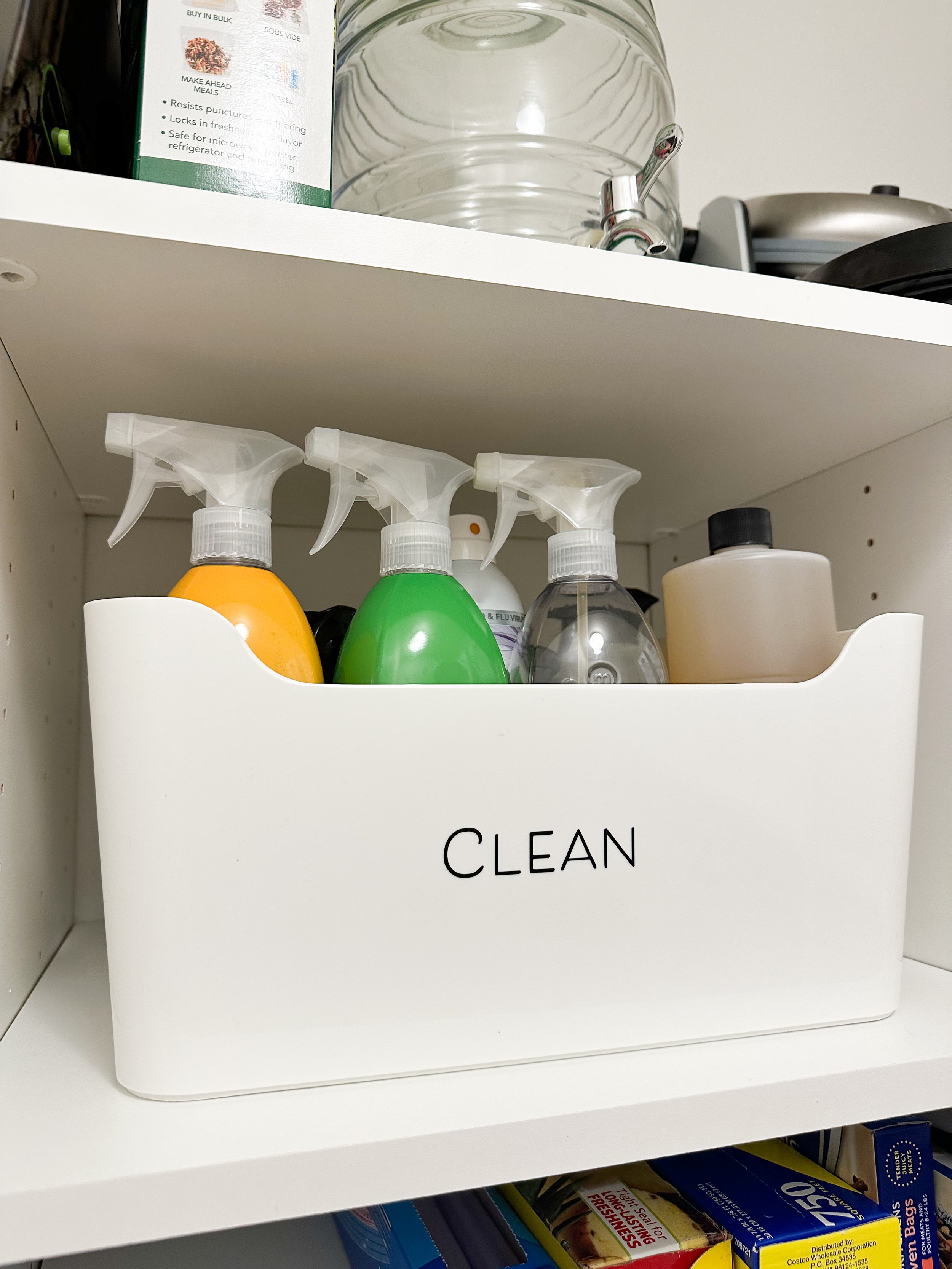 A white storage container labeled 'CLEAN' holding spray bottles and cleaning supplies on a kitchen shelf.