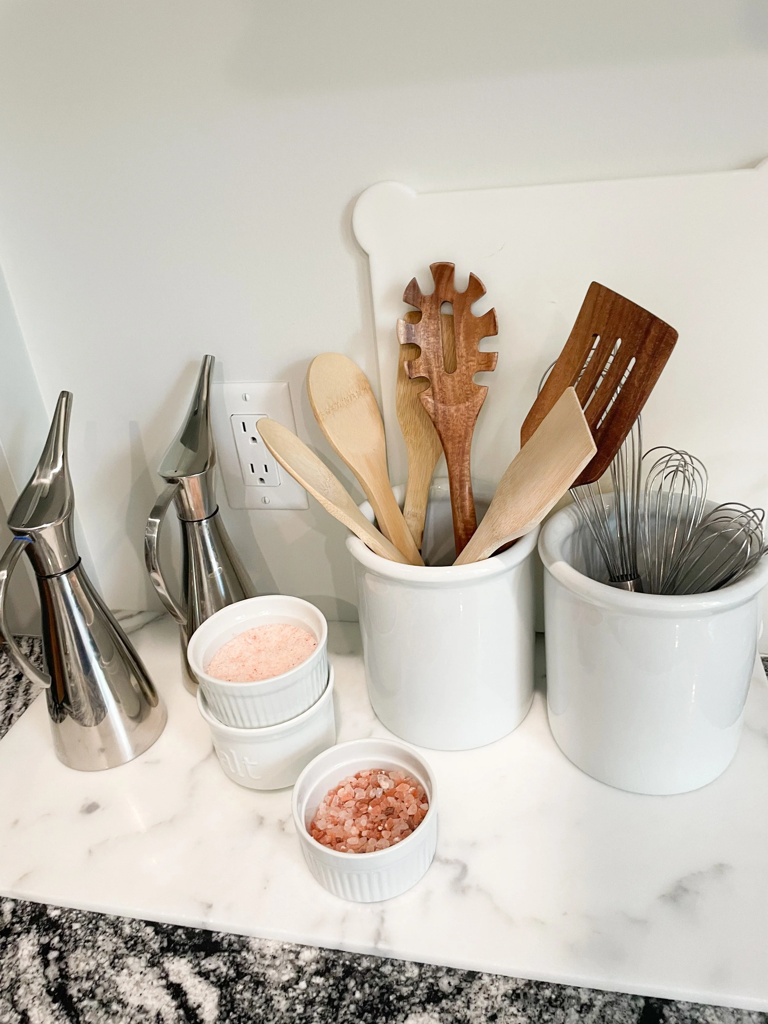 Kitchen counter with salt and pepper shakers, two small bowls of pink Himalayan salt and pink salt, and white ceramic containers holding wooden and metal kitchen utensils and whisks.