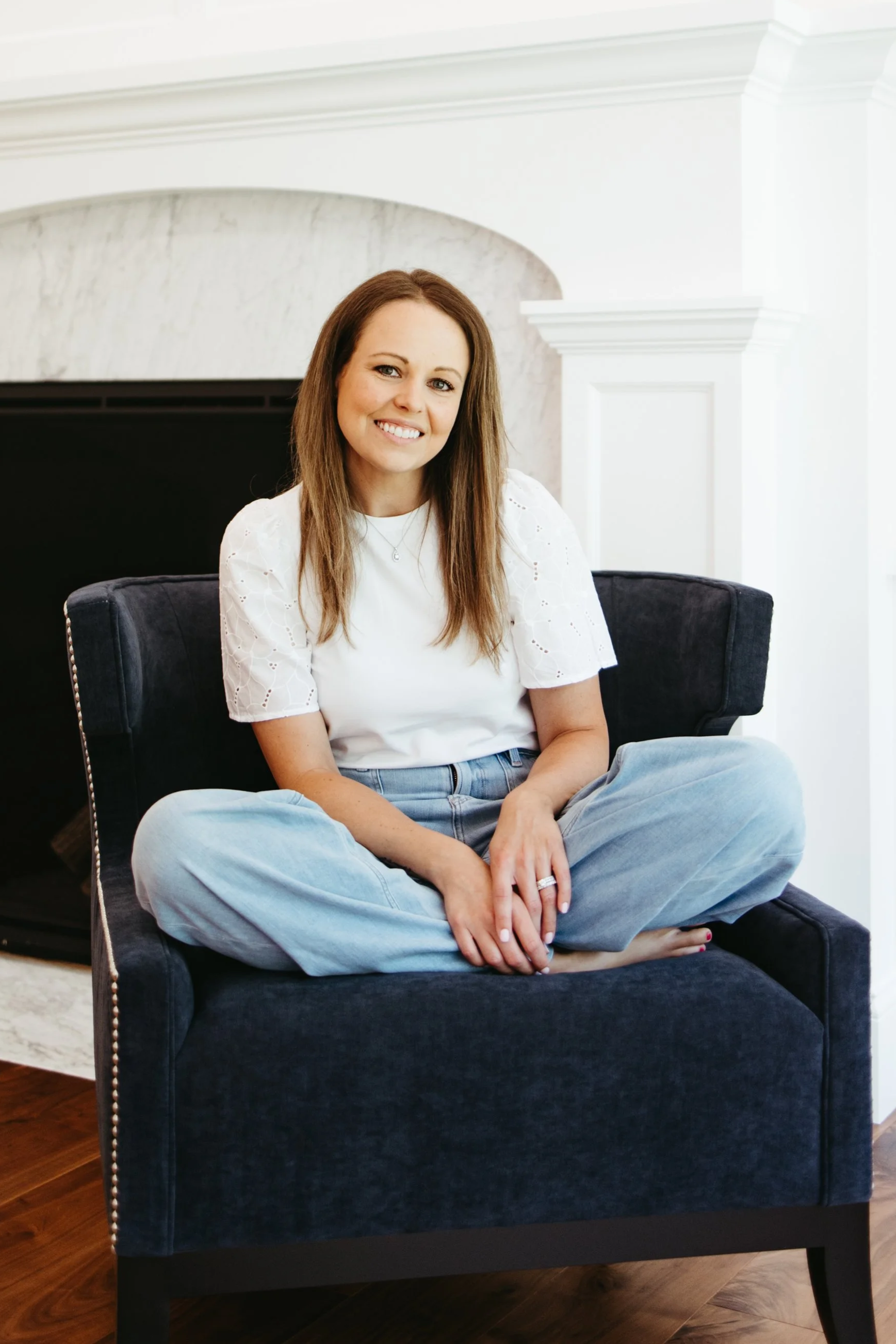 A woman with brown hair, wearing a white eyelet blouse and light blue jeans, sitting cross-legged on a navy blue chair in a bright room with a white wall and a fireplace in the background.
