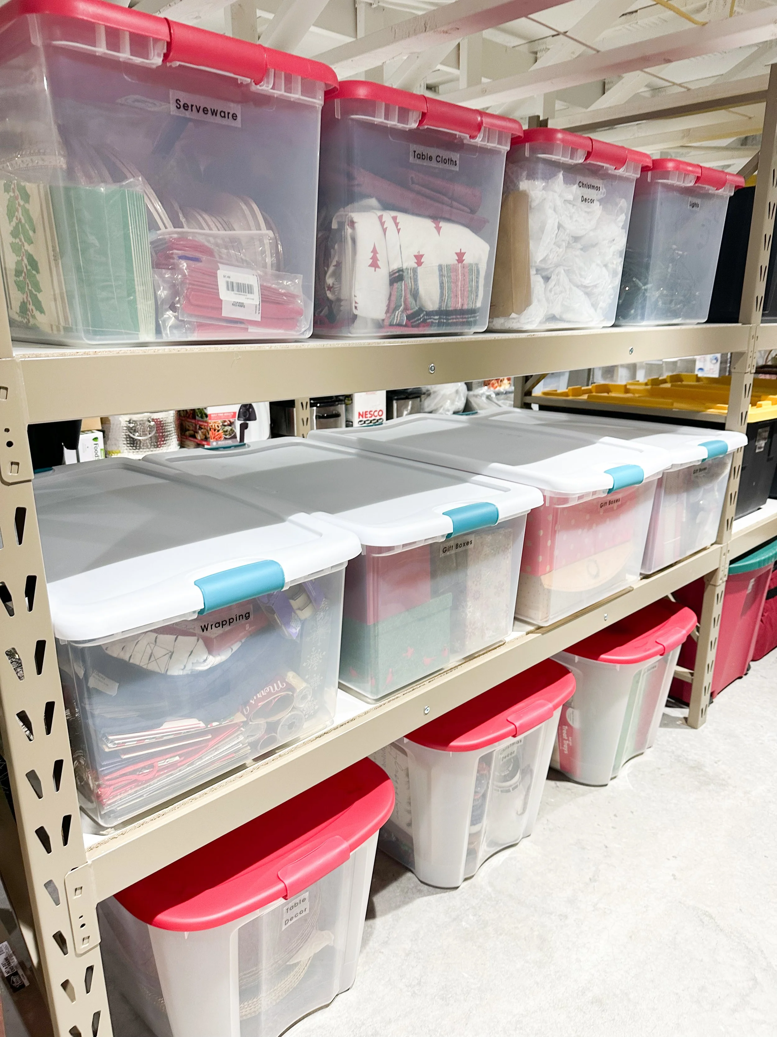 Shelving unit with clear plastic storage bins labeled for holiday and household decor, including 'Serveware,' 'Table Clothes,' 'Christmas Decor,' 'Lights,' 'Wrapping,' and 'Gift Boxes.'