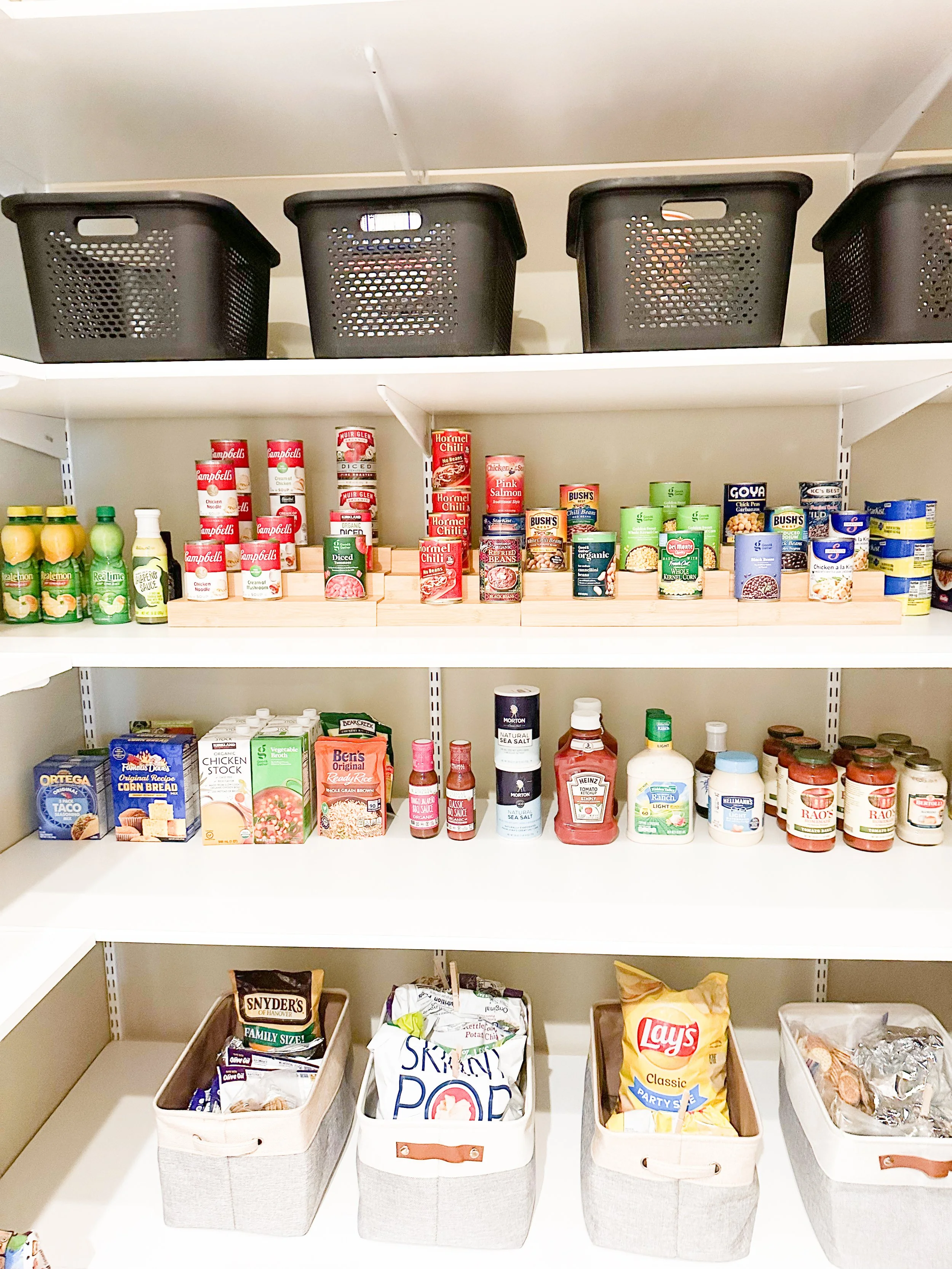 Organization pantry shelf with black baskets on top, canned goods, boxed foods, jars, and snack bags.
