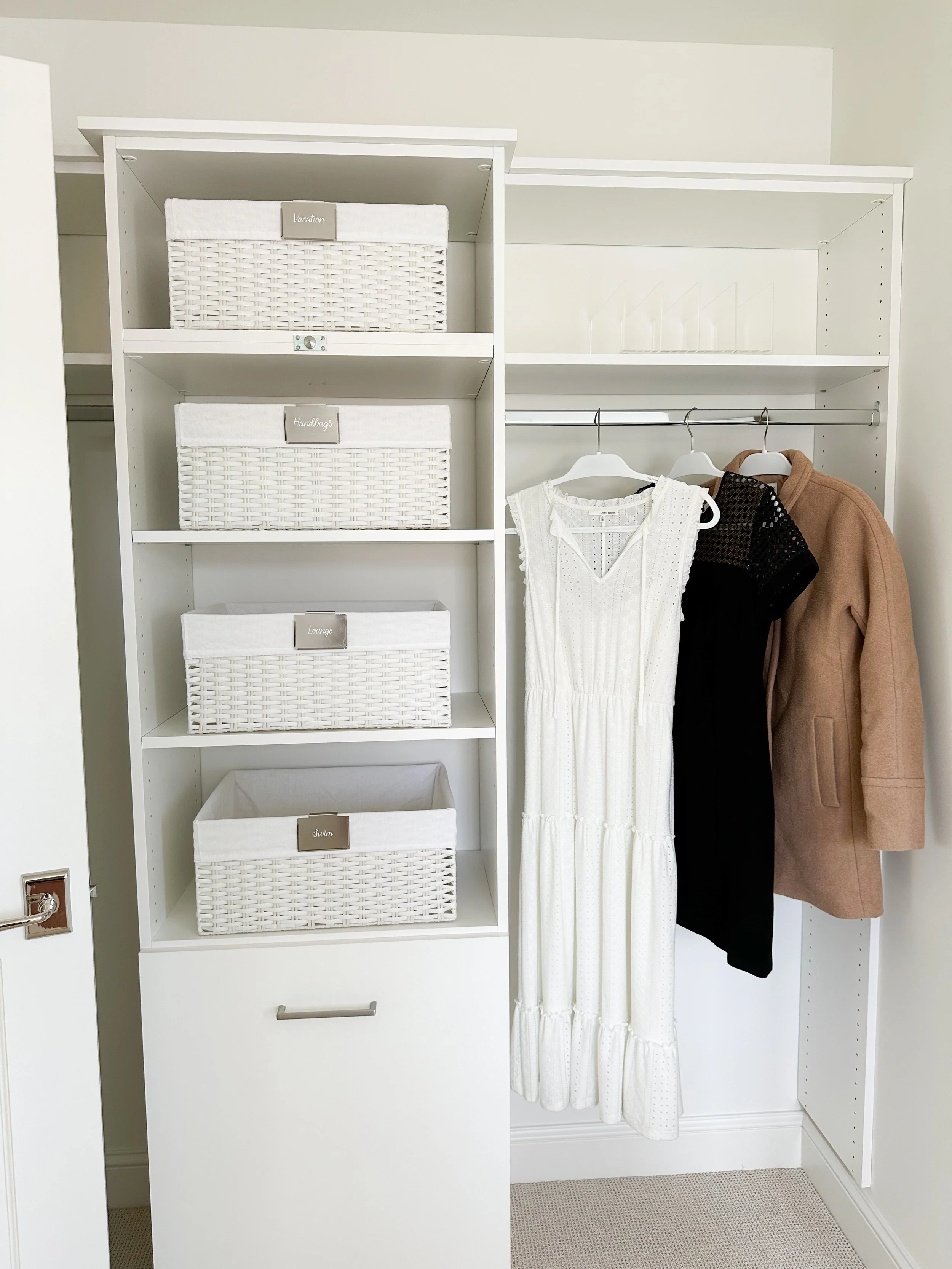 A white closet organizing system with labeled white baskets on the left shelves and hanging clothes, including a white dress, a black dress, and a tan coat, on the right.