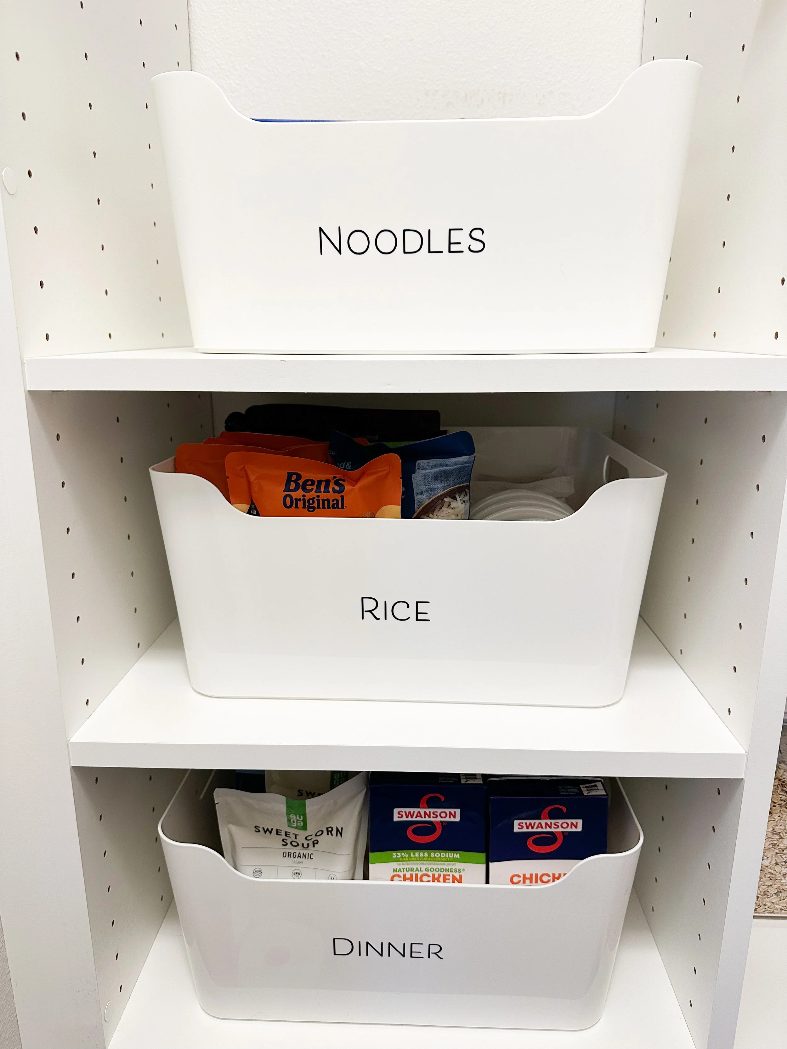 White storage bins labeled 'NOODLES', 'RICE', and 'DINNER' on a white shelving unit filled with food items