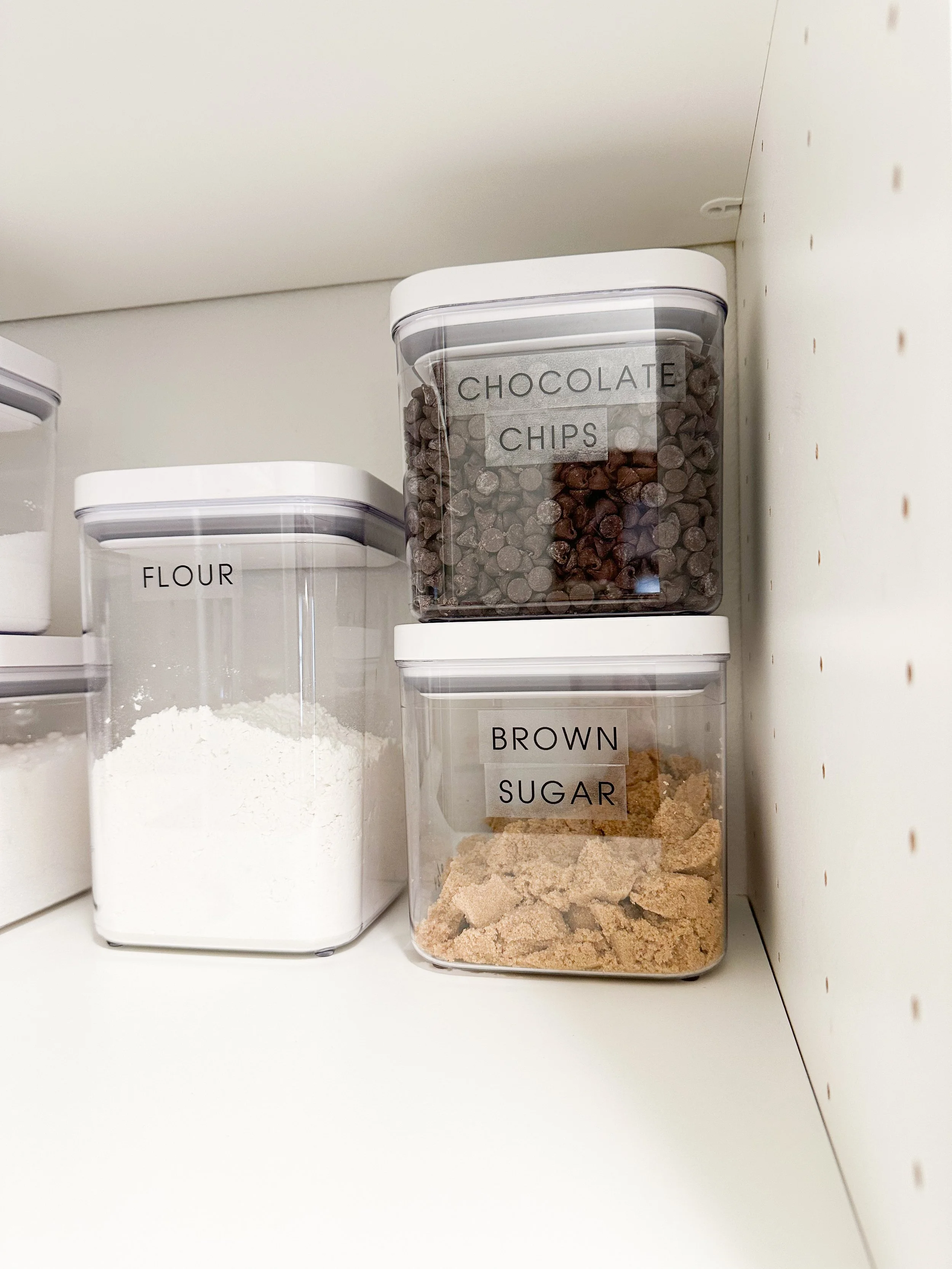 Clear pantry containers labeled 'flour,' 'chocolate chips,' and 'brown sugar' on a white shelf.