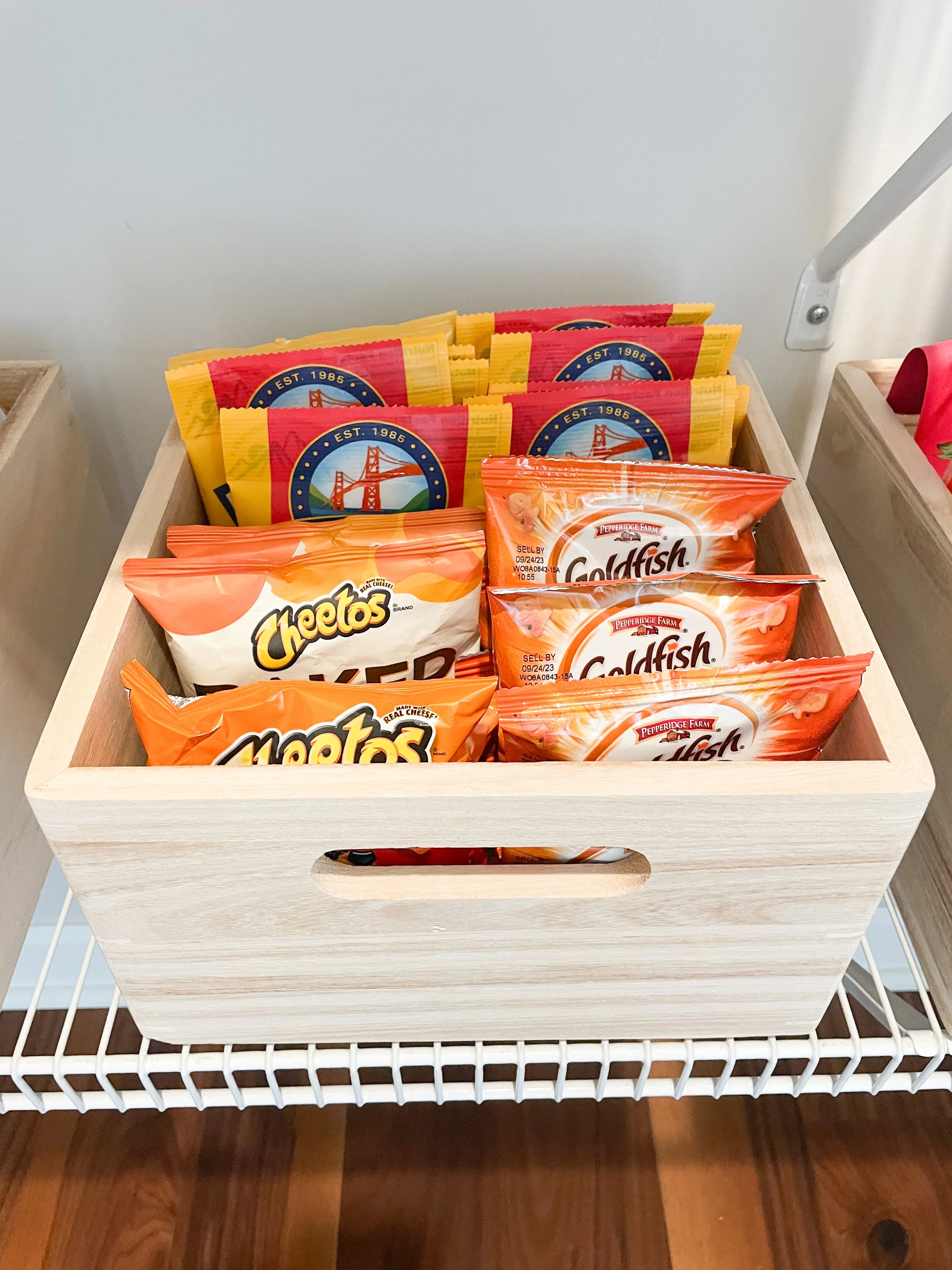 A wooden box containing packets of Los Angeles-themed souvenirs, Cheetos cheese snacks, and Pepperidge Farm Goldfish crackers, placed on a white wire shelf.