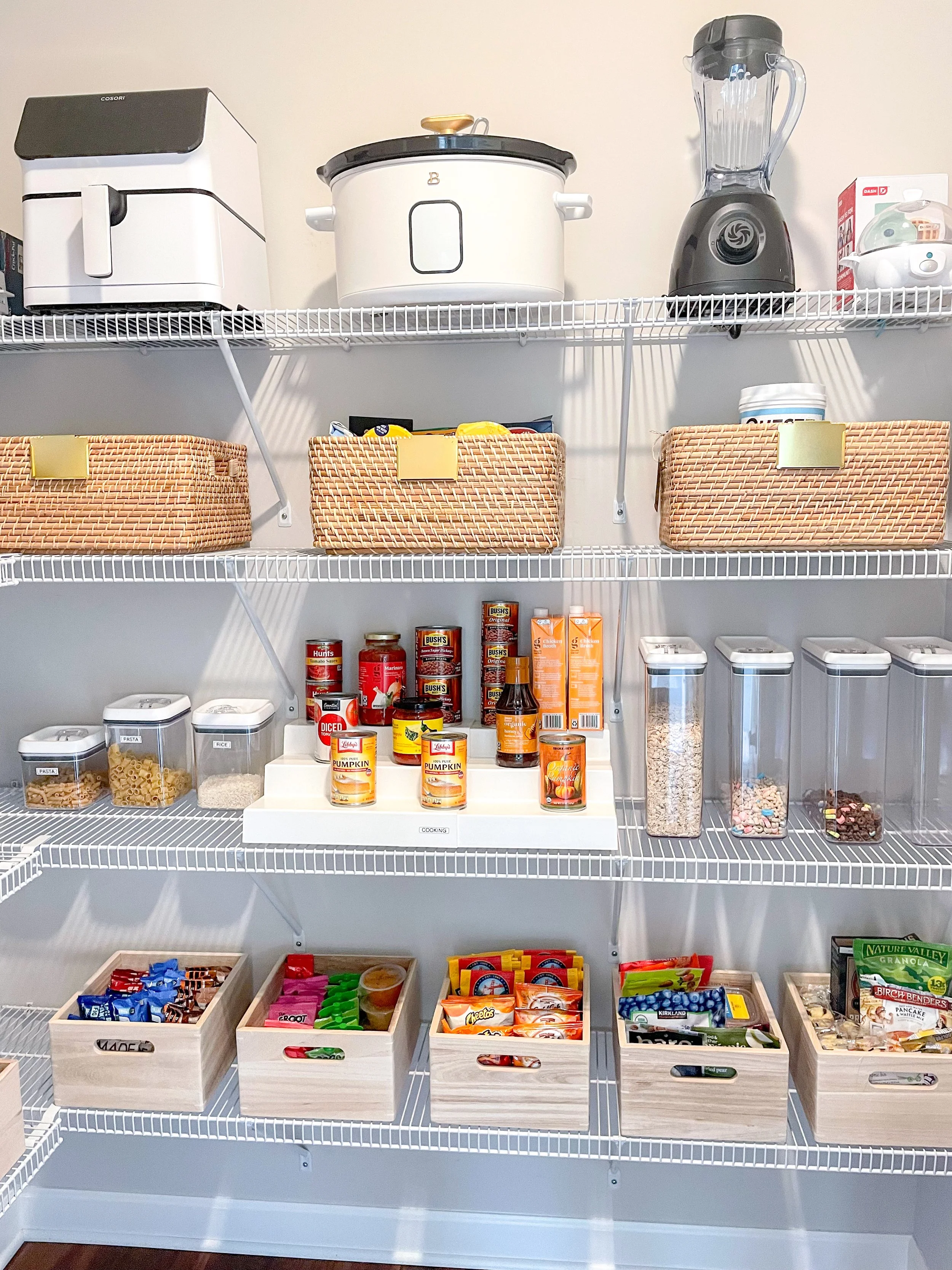Organized pantry with wire shelves holding kitchen appliances, baskets, canned goods, snacks, and cereals.