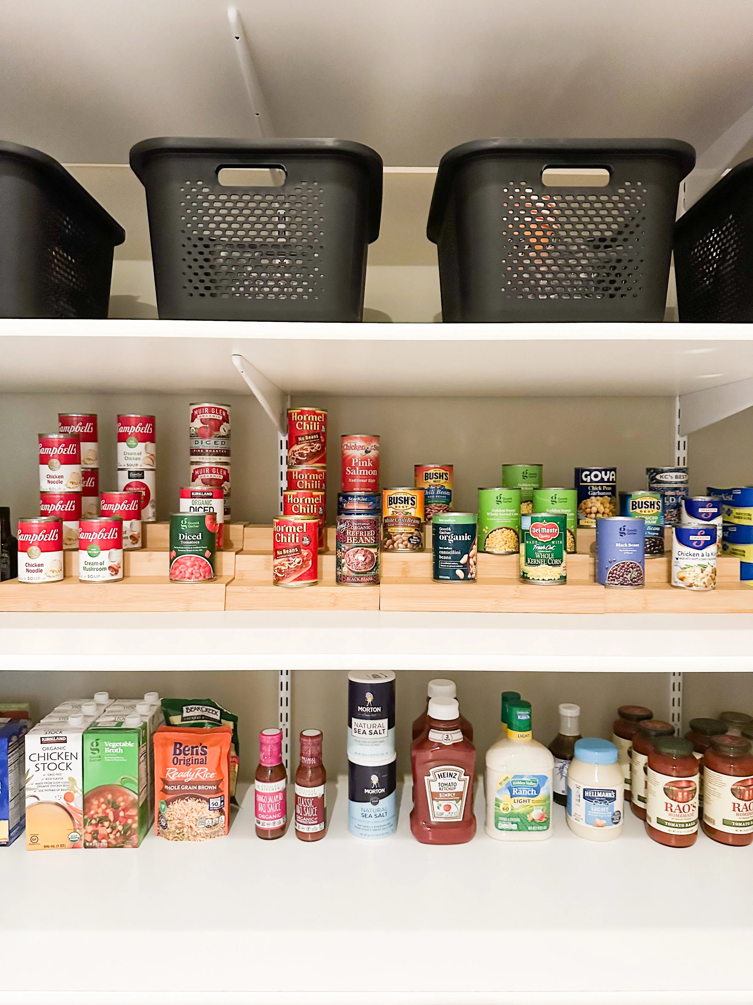 Pantry shelf filled with canned soups, beans, vegetables, and condiments, arranged neatly with black baskets on the top shelf.