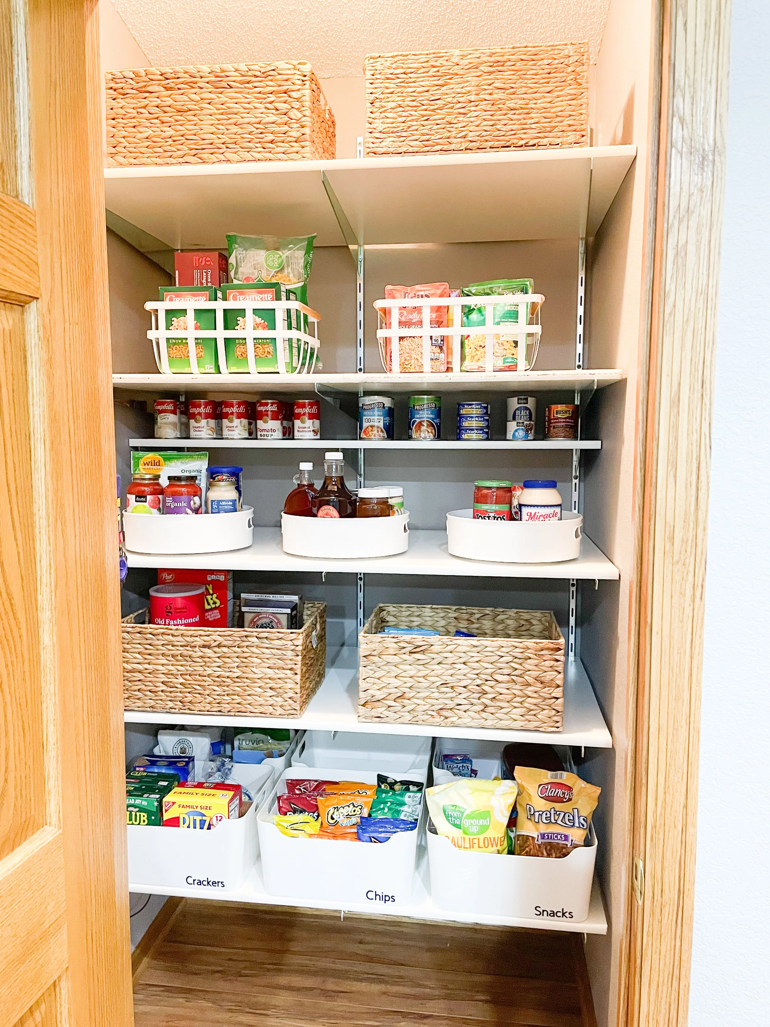 A pantry with multiple shelves containing canned goods, snacks, chips, cookies, and other food items organized in white baskets and woven containers.