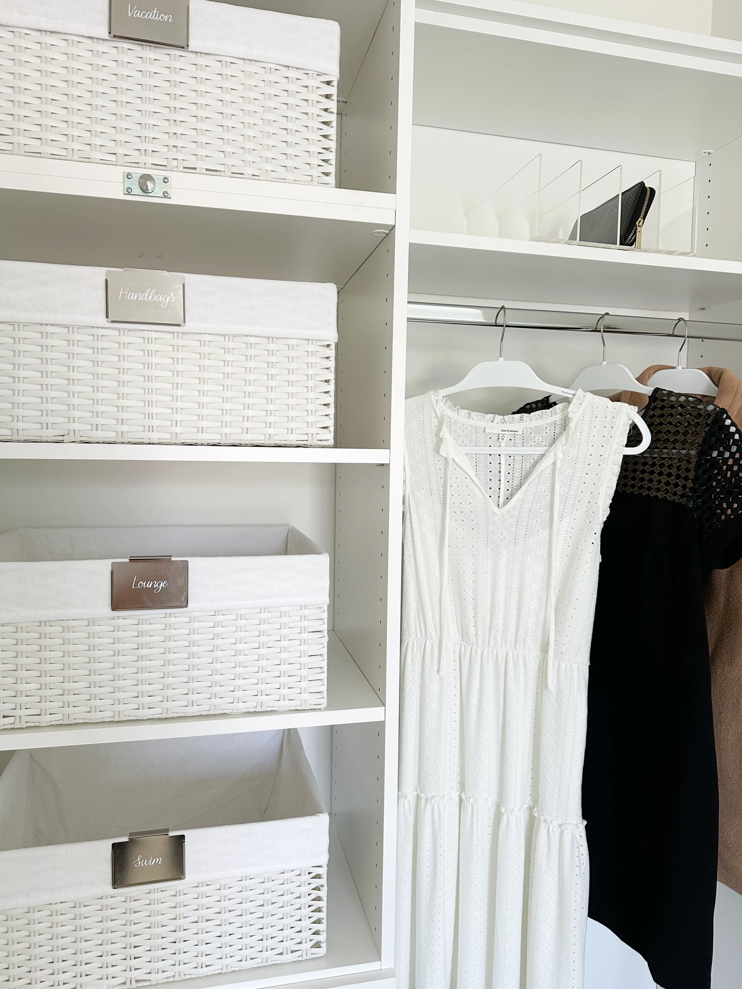 White shelves with labeled storage bins, clothing hanging on hangers including a white dress and a black dress.