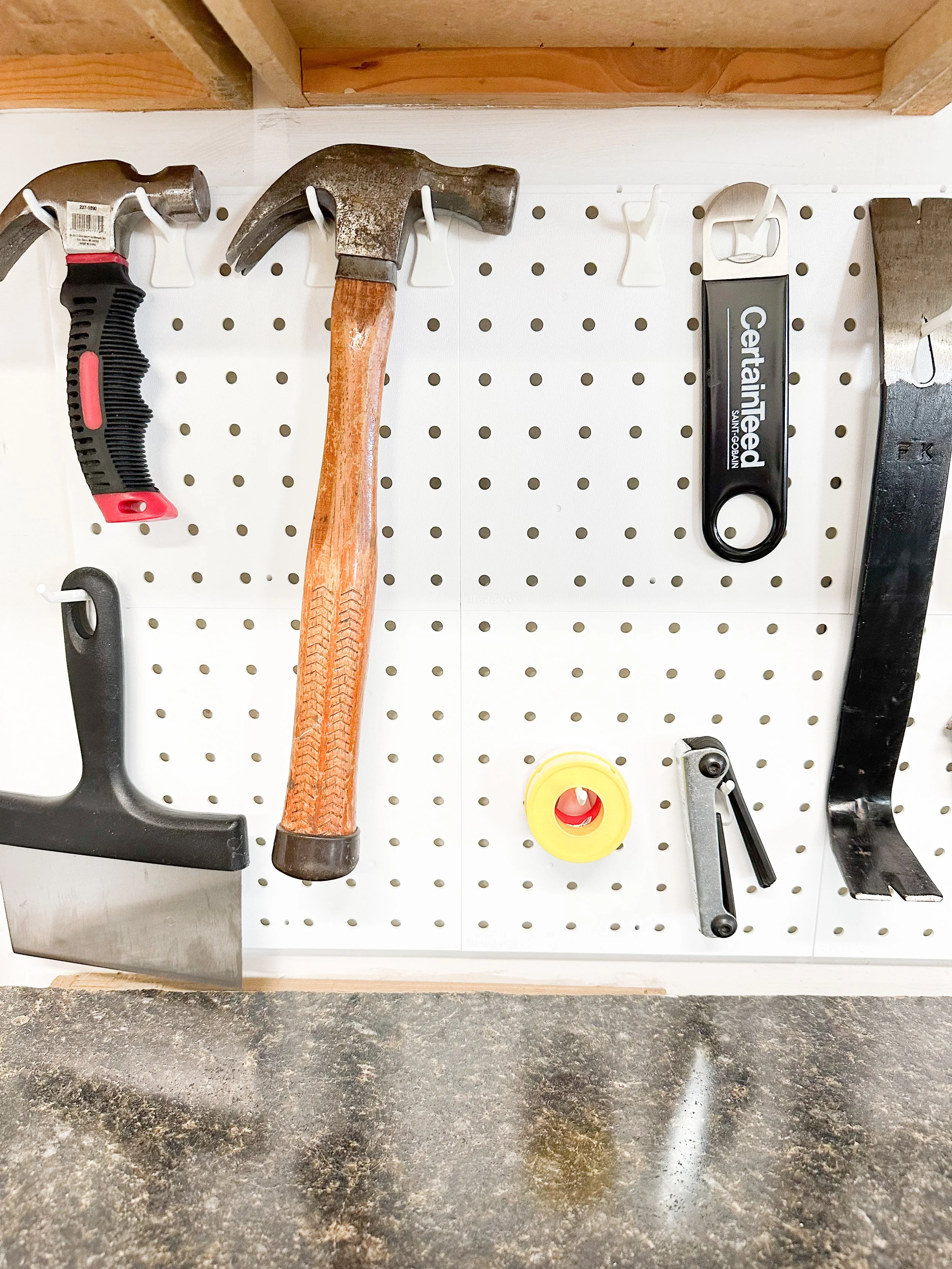 Pegboard with tools including a hammer, a hand saw, a scraper, a tape measure, and a tool handle, mounted on a wall above a concrete surface.