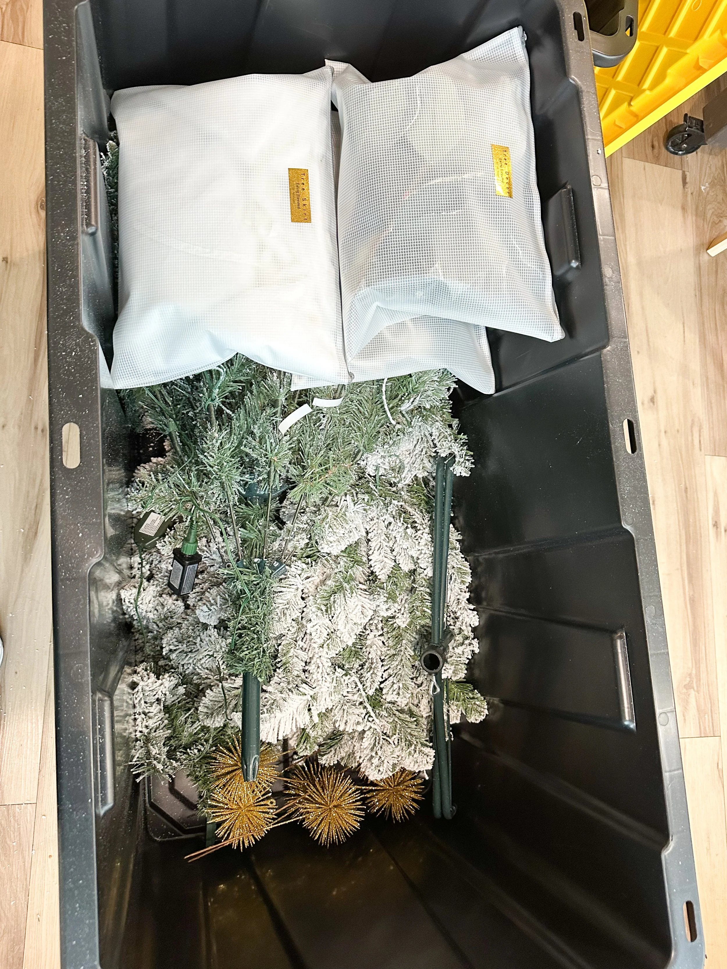 A Christmas tree with snow-like flocking and gold ornaments, packed inside a large black storage container with two pillows and some disassembled parts.