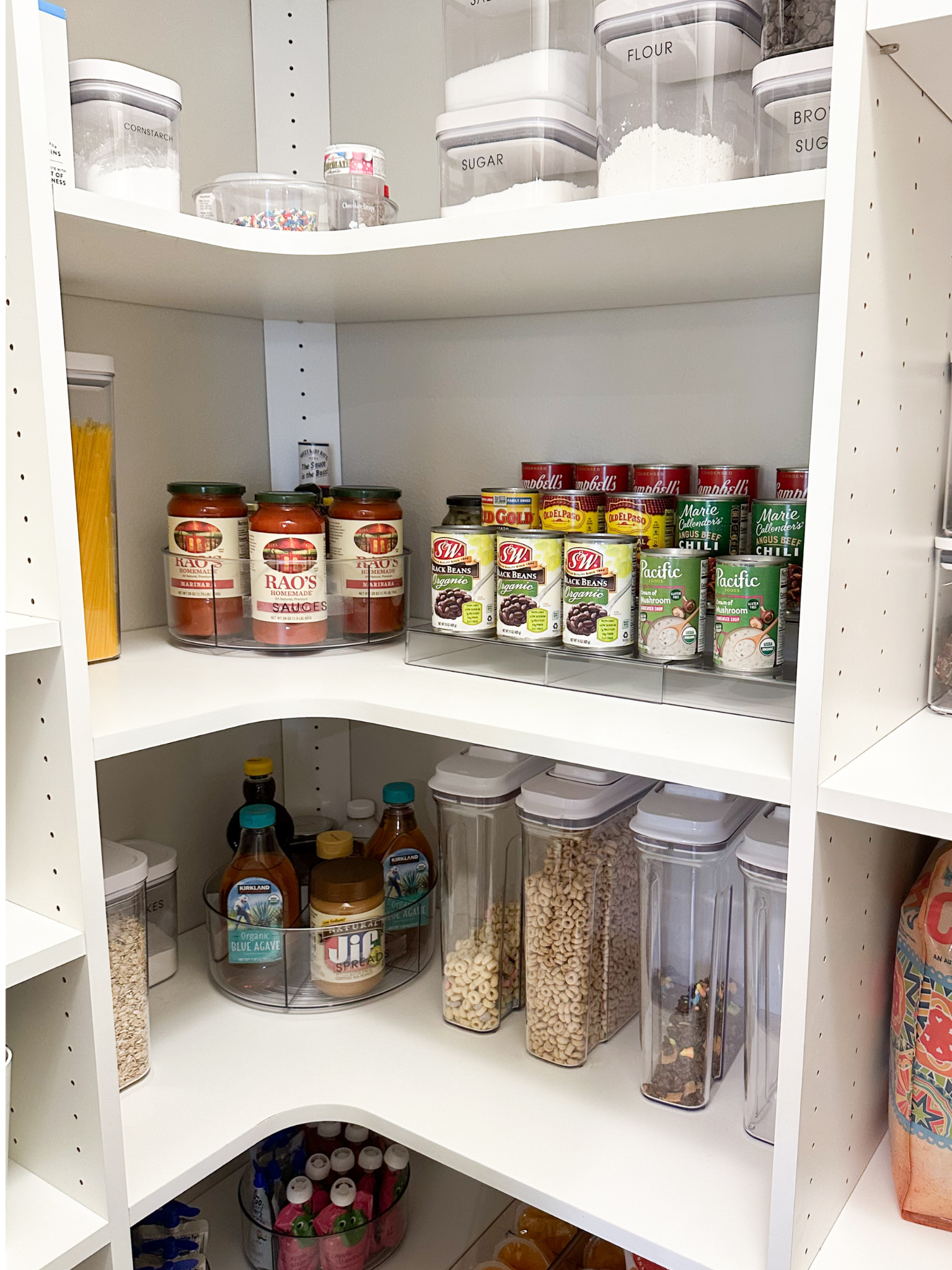 Pantry shelf containing canned black beans, soup, sauces, and other food items stored in clear containers.
