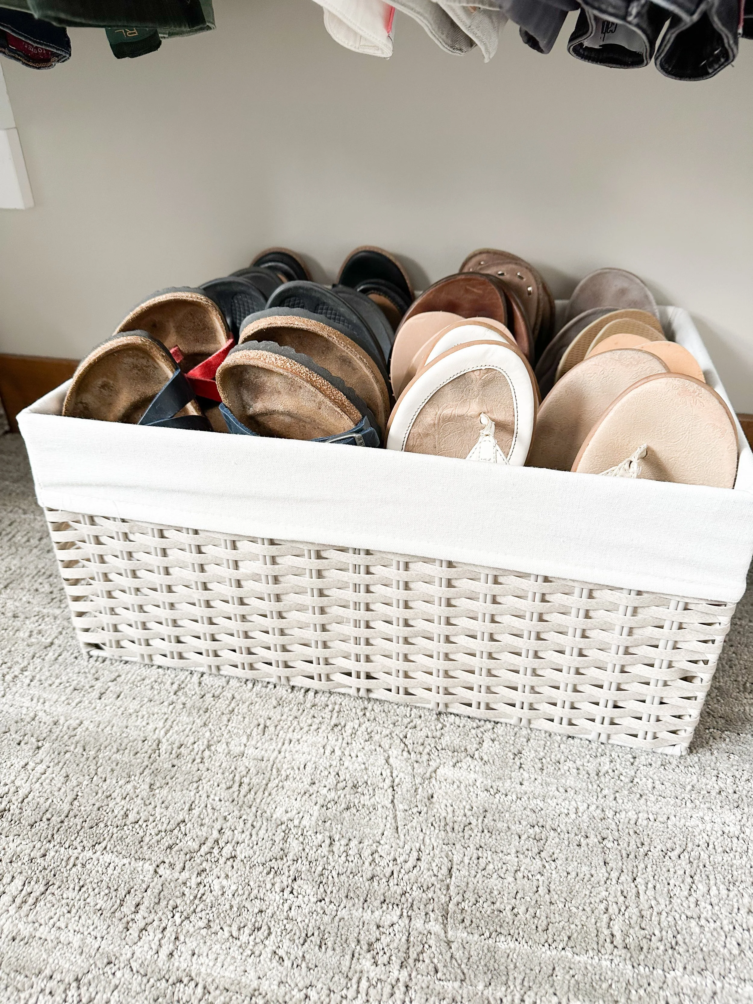 A white woven basket filled with various pairs of slide sandals in neutral tones, placed on a beige carpet.