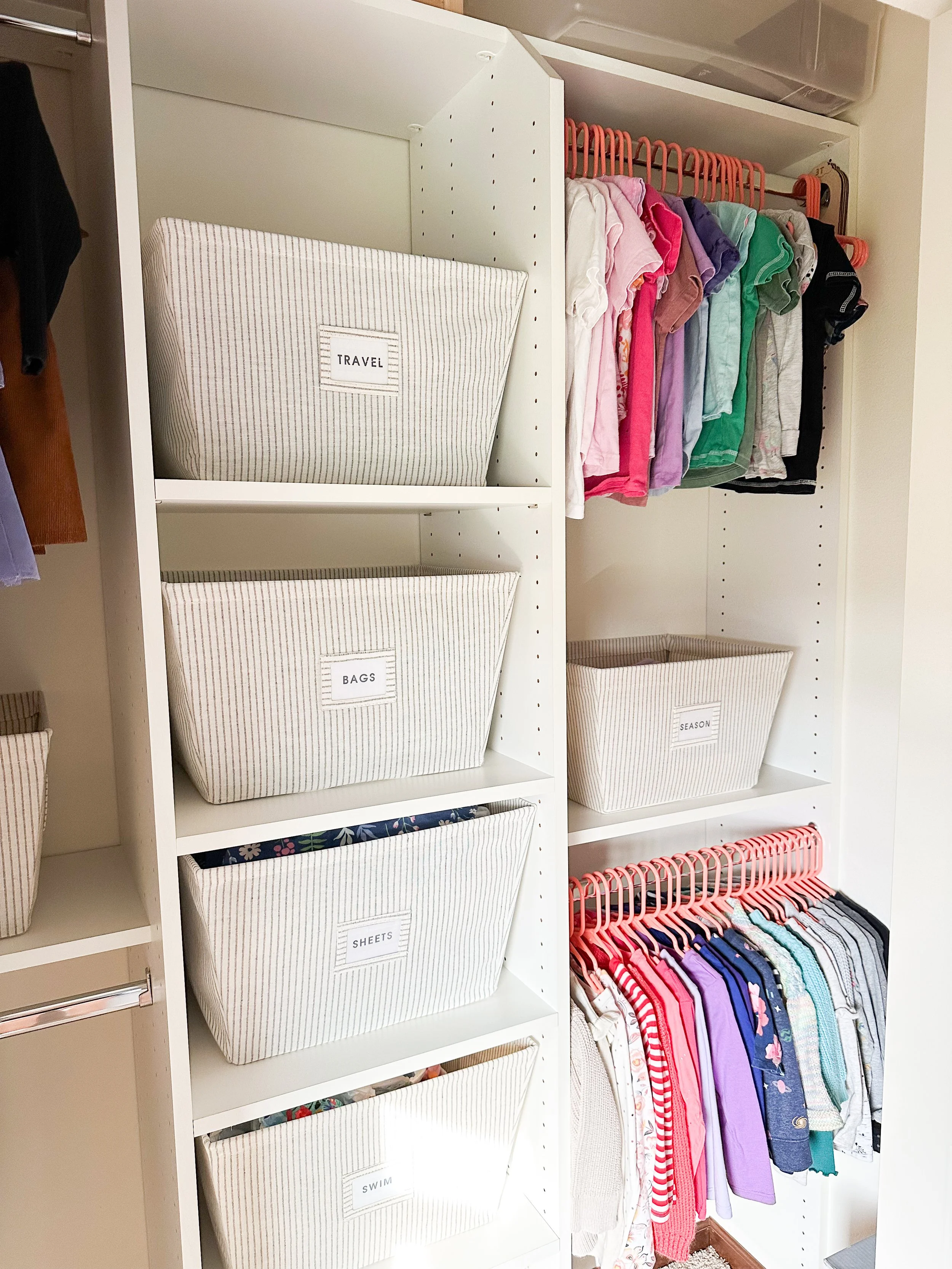 A closet with white shelves holding labeled striped fabric bins for travel, bags, sheets, and swim. Above, a clothing rod with colorful children's clothes on pink hangers, and below, a rod with assorted children's shirts and tops.