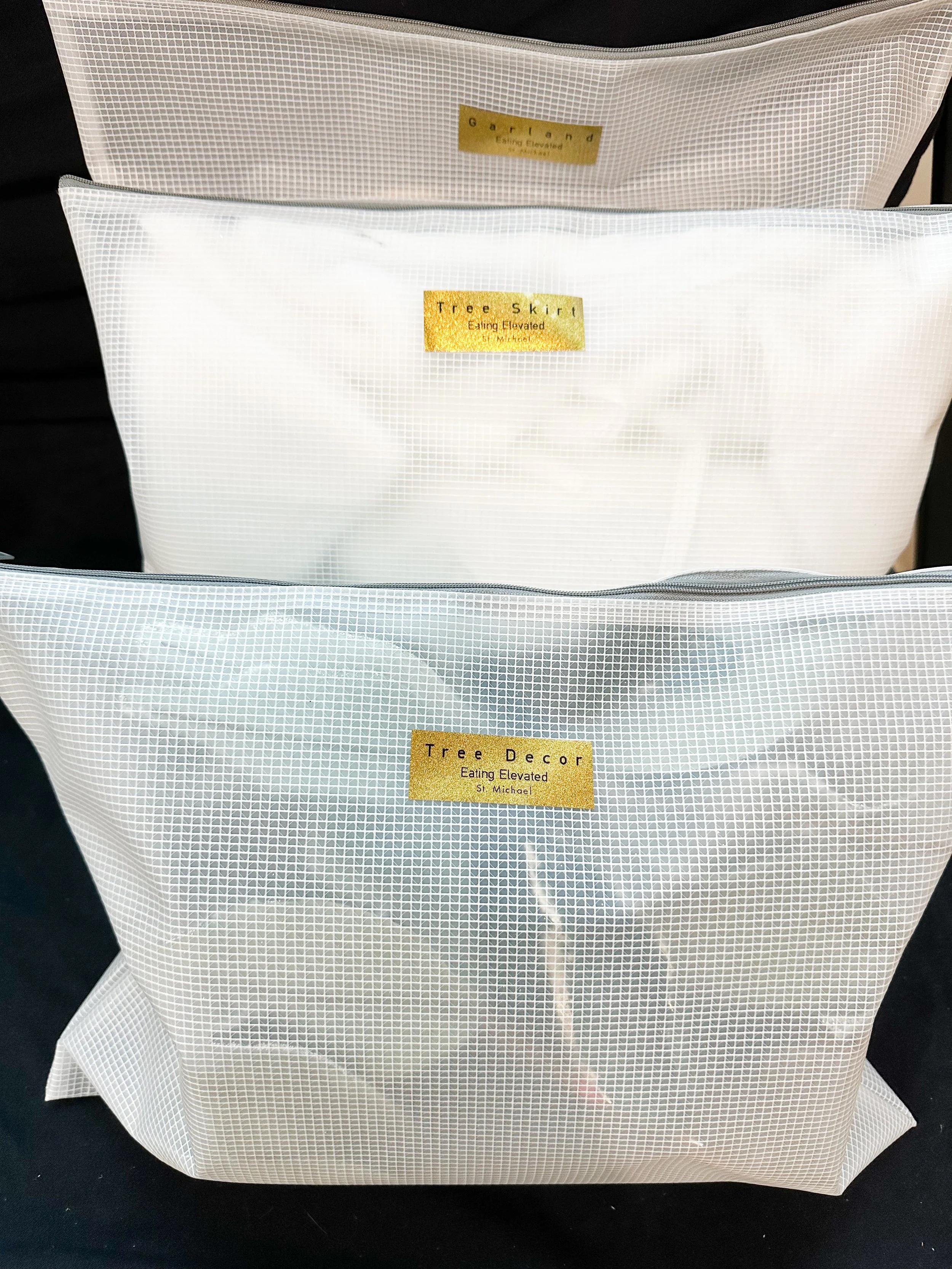 Three white mesh bags with gold labels that read 'Garland,' 'Tree Skirt,' and 'Tree Decor,' each containing Christmas decorations or ornaments.