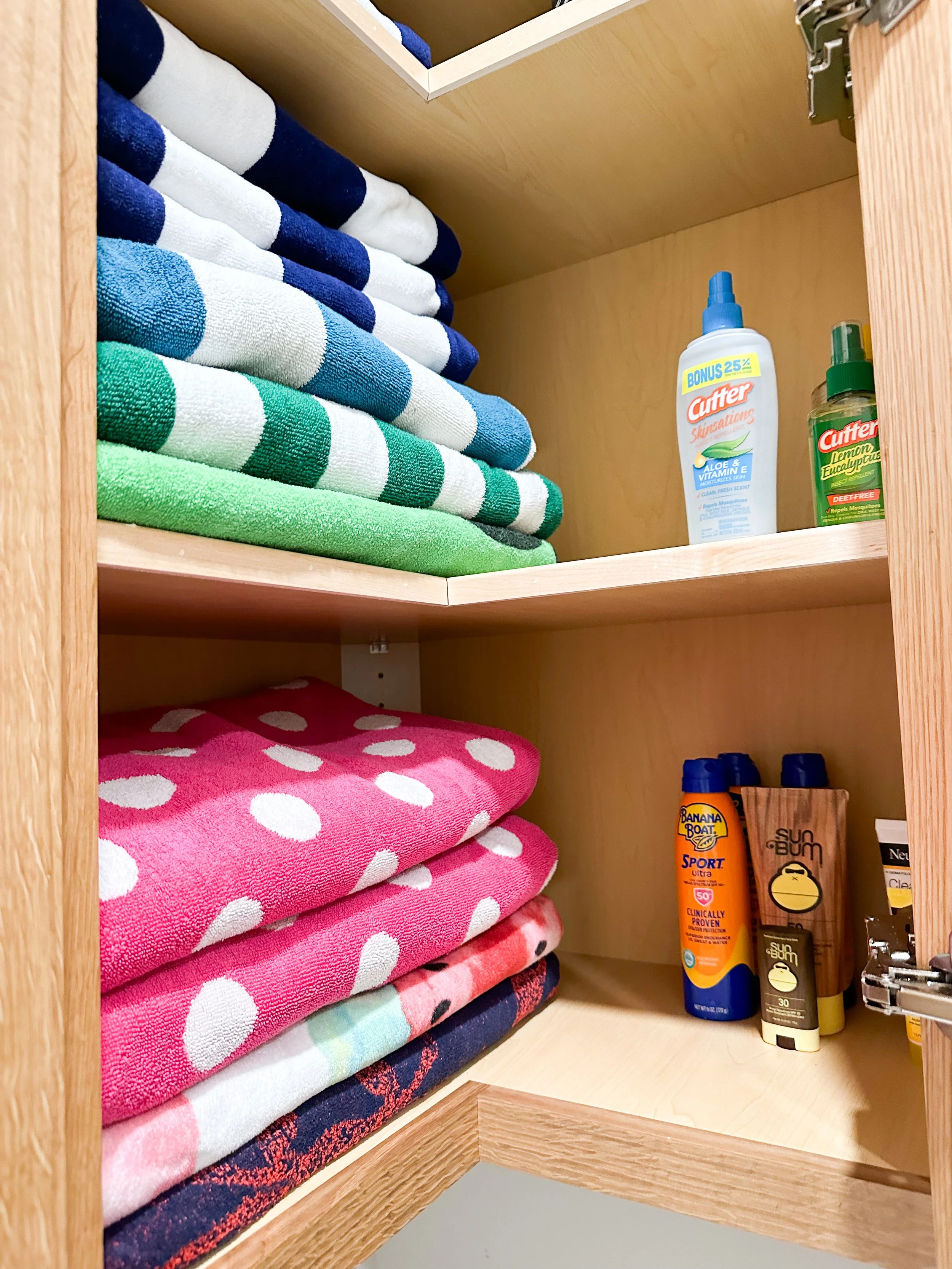 A wooden cabinet with neatly folded towels in blue, green, pink, and patterned designs, and bottles of sunscreen and insect repellent on the shelves.
