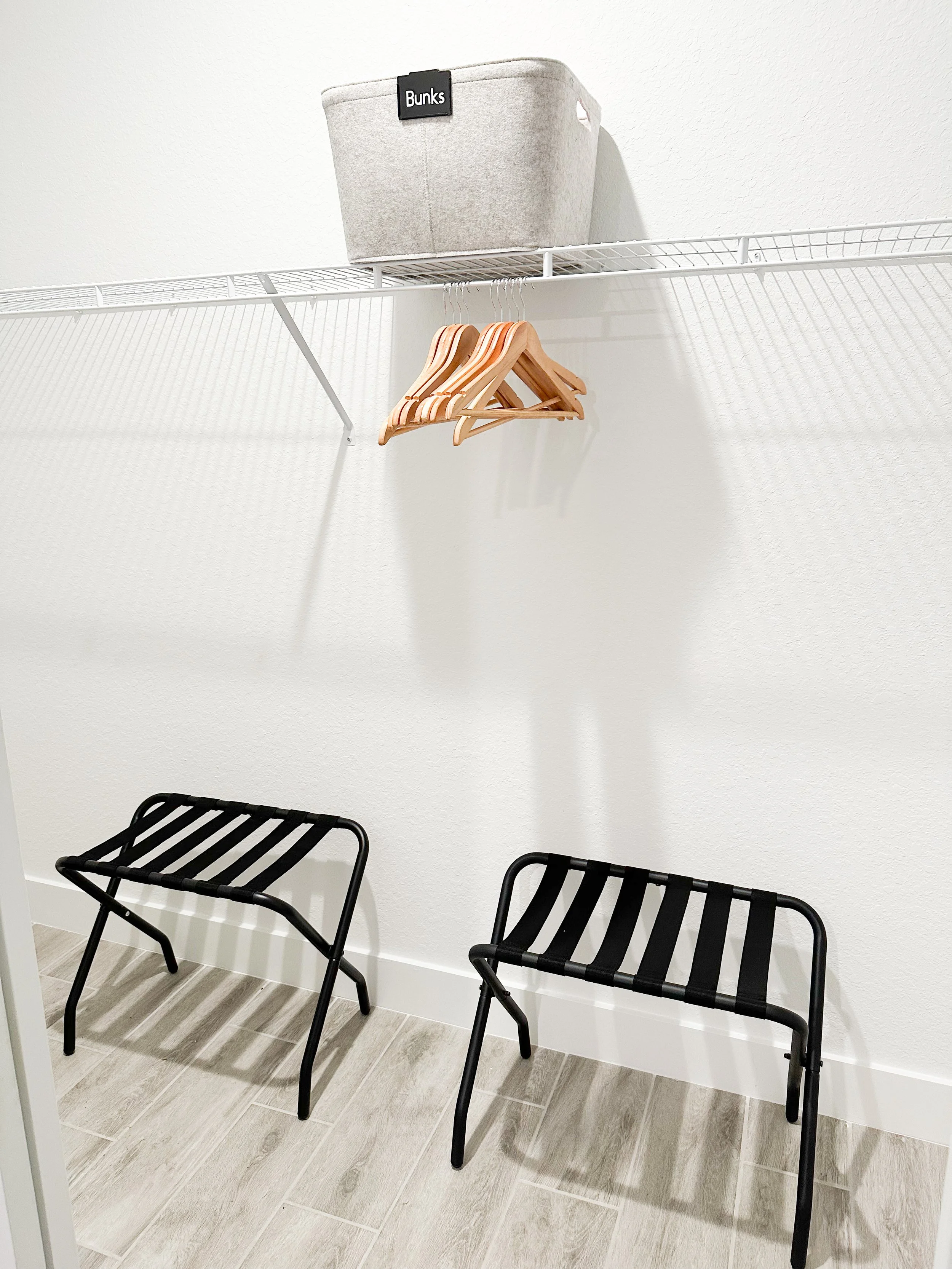 A minimalist closet with a white wire shelf holding a storage bin labeled 'Bunks' and a few wooden hangers. Below, two black metal benches with slatted seats are positioned on a gray wood-look tile floor.